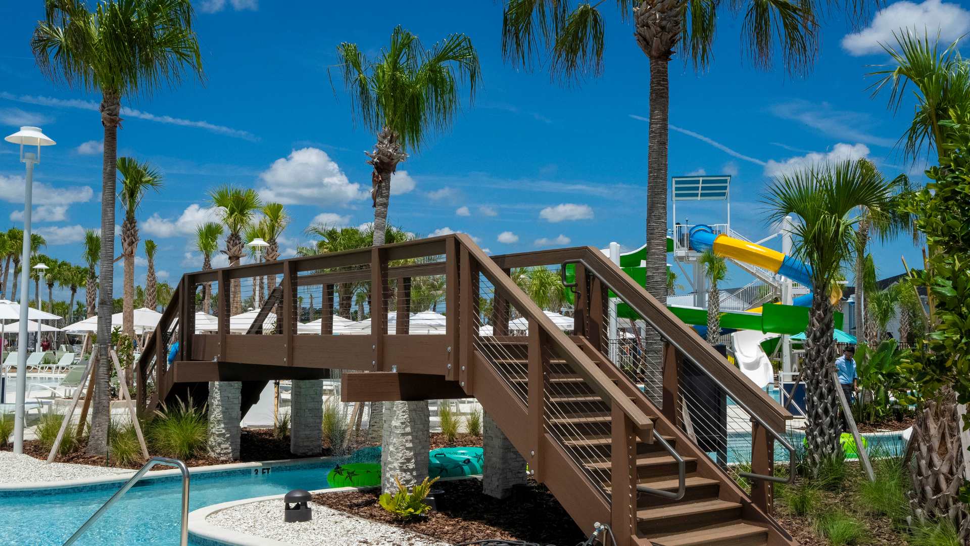 Sturdy wooden bridge with stairs and cable railings spanning over the lazy river, providing access across the communal pool area, surrounded by lush landscaping and palm trees.