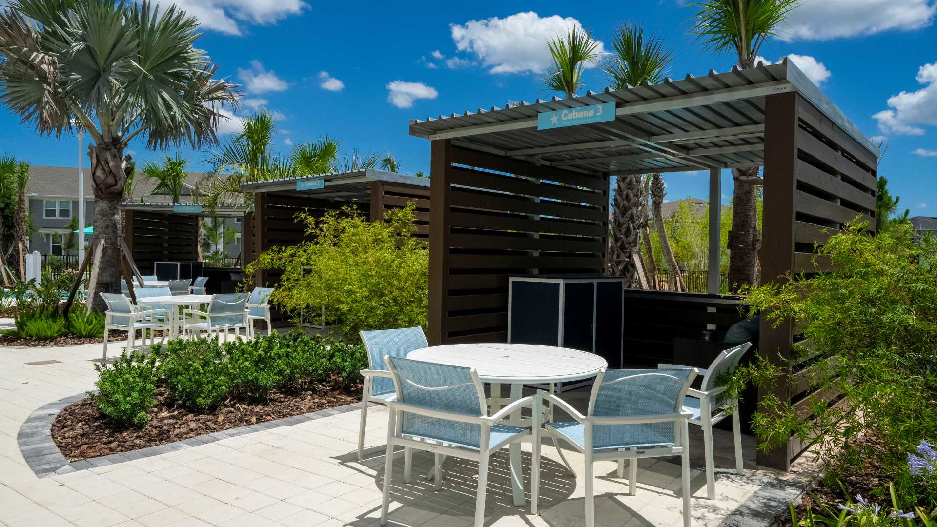 Inviting outdoor cabanas with slatted roofs and privacy screens, each furnished with a white round table and light blue chairs, set on a paved patio amidst lush green foliage.