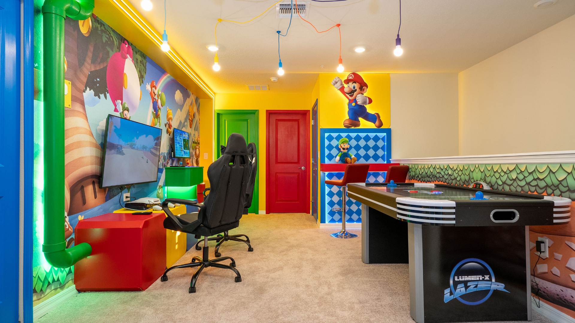 Vibrant Mario-themed game room with a large TV for gaming, a black gaming chair, and an air hockey table, all set against colorful murals and bright yellow walls. Exposed light bulbs hang from red and yellow cords, adding to the playful atmosphere.
