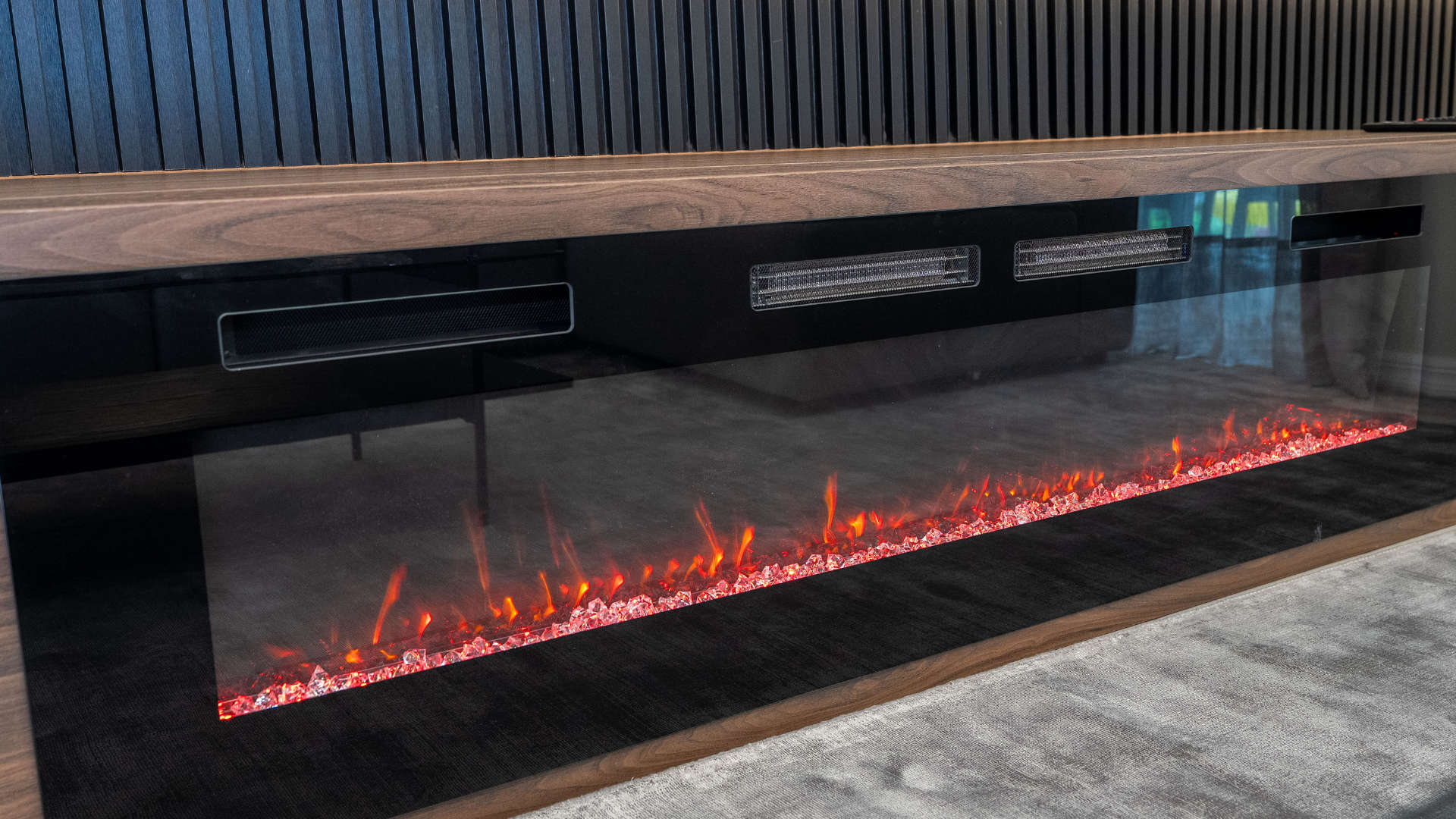 Detailed view of the modern electric fireplace, showcasing its glowing red and orange crystal-like embers within a sleek black and wood-framed unit. The fireplace is integrated into a larger entertainment wall with dark slatted accents.