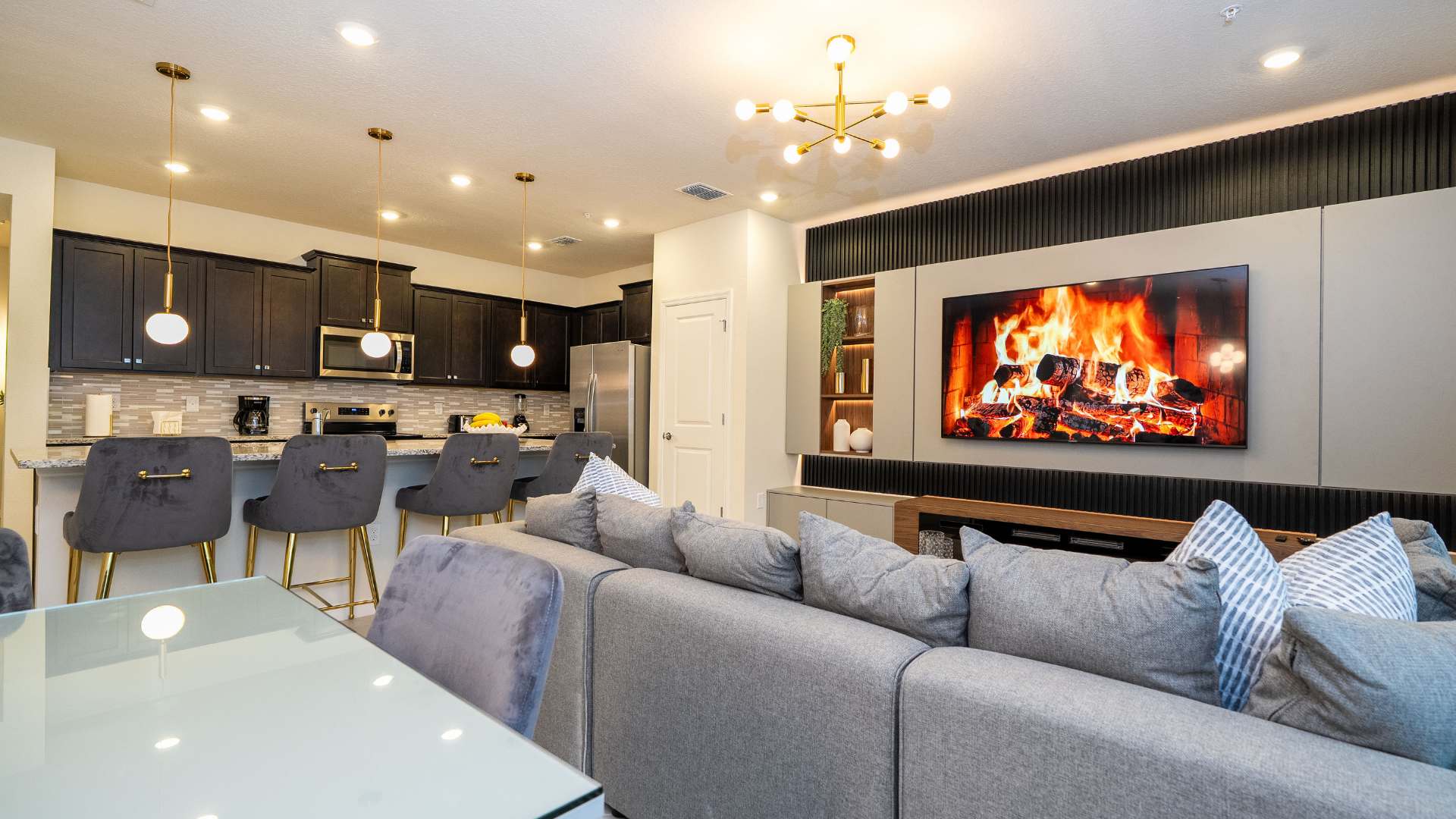 Open-concept view of the living room and adjacent kitchen, showing the grey sectional sofa facing the entertainment unit with a TV and electric fireplace. The kitchen features dark cabinetry, stainless steel appliances, and a breakfast bar with grey velvet stools.