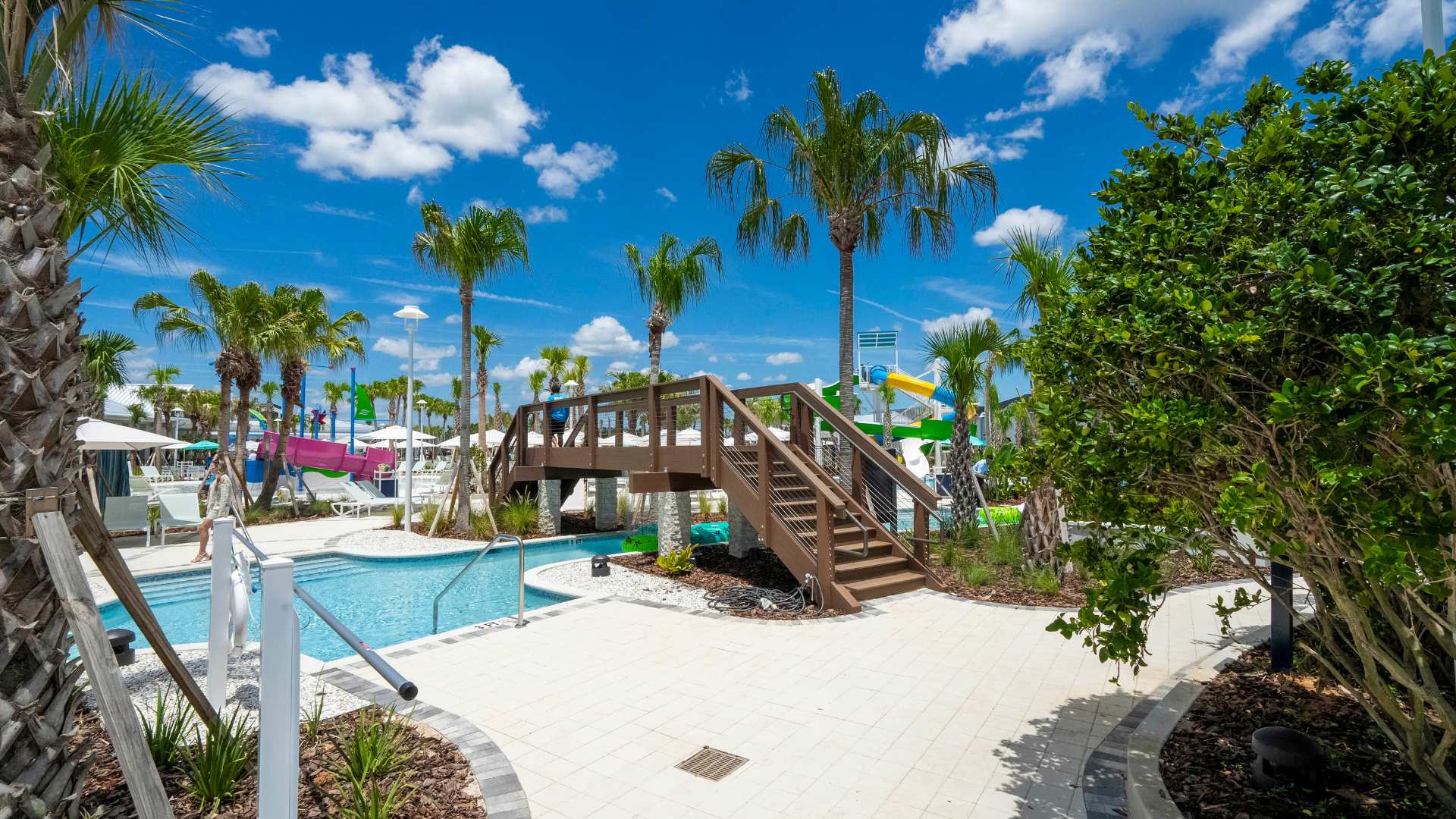Scenic view of the communal pool area featuring a wooden bridge over the lazy river, extensive paved walkways, and tropical landscaping with palm trees under a bright blue sky.