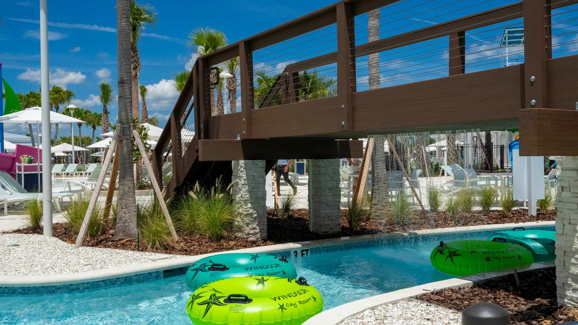 A winding lazy river with bright green and blue inner tubes floating in clear blue water, shaded by a modern wooden bridge and surrounded by lush palm trees and white gravel landscaping.