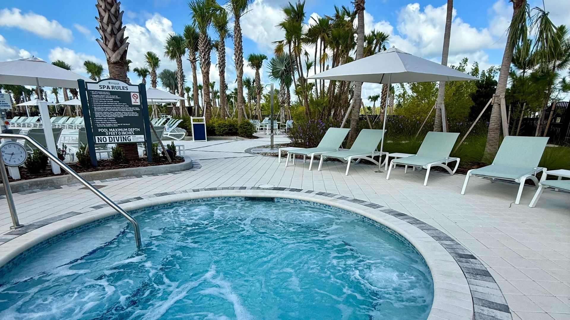 Circular outdoor hot tub with bubbling water, surrounded by light-colored pavers, several light green lounge chairs, and lush palm trees under a partly cloudy sky.