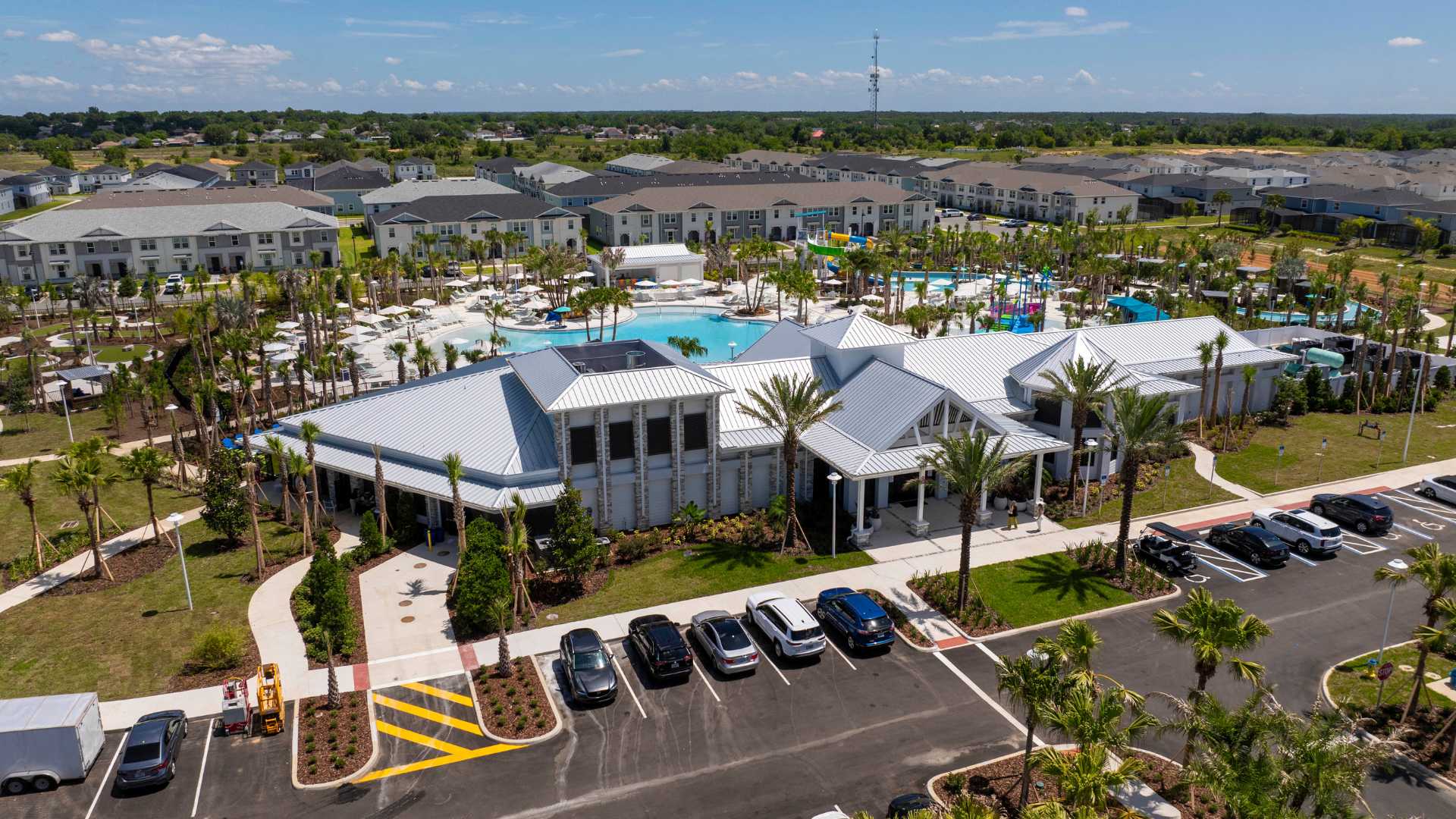 Aerial view of a large, modern resort clubhouse with a light-colored exterior and multiple gabled metal roofs, surrounded by palm trees, a parking lot, and a sprawling pool complex in the distance.