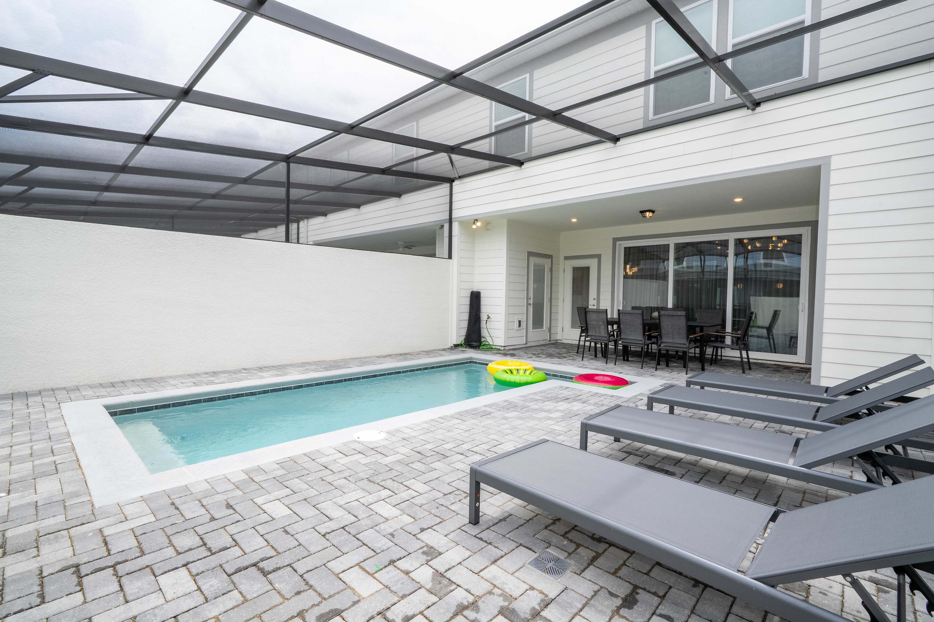 A private swimming pool with clear blue water and colorful inflatable rings is set within a gray brick-paved patio, enclosed by a screen cage. Four modern gray lounge chairs are neatly arranged along a white wall, offering a sunny spot for relaxation.