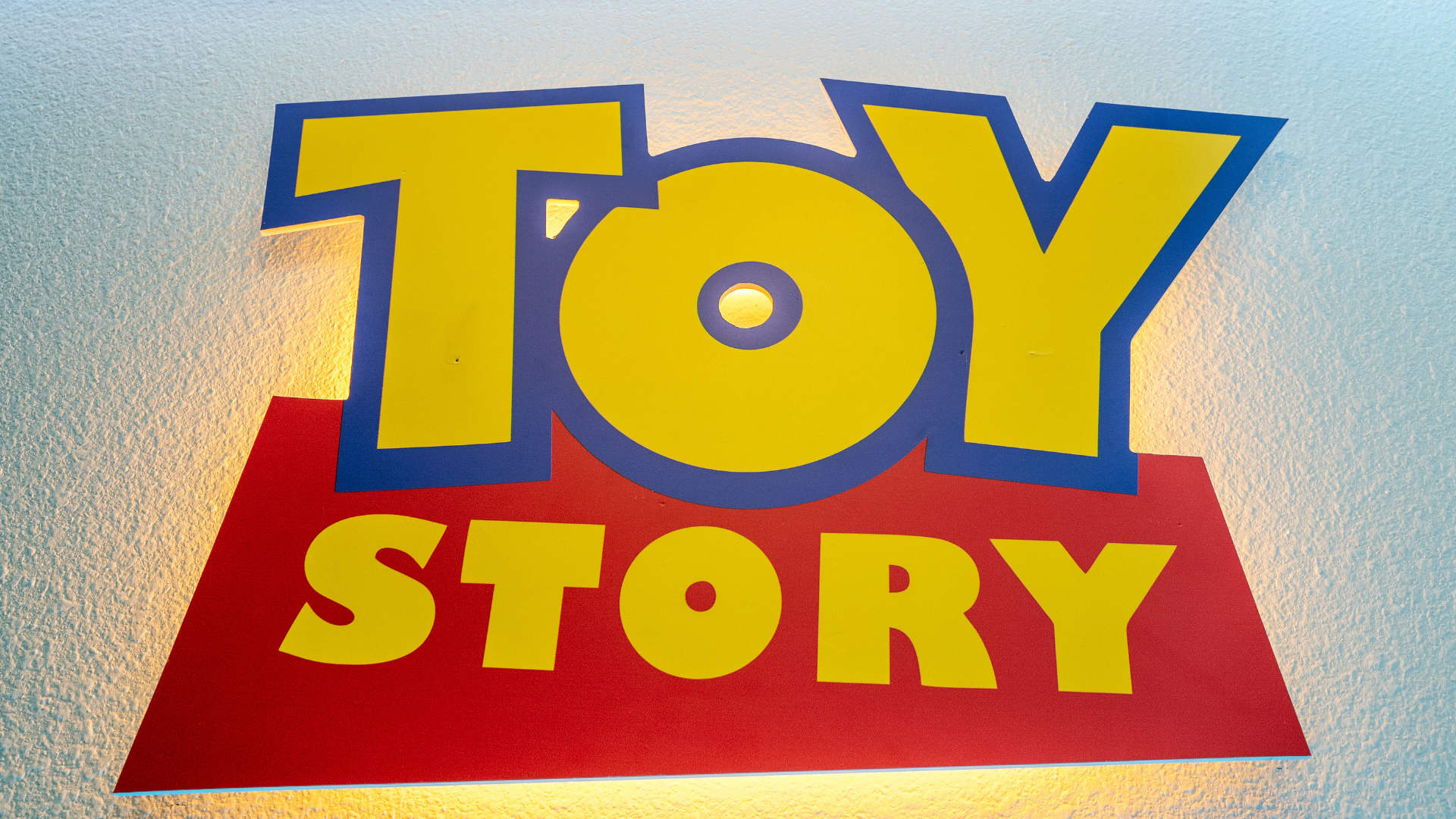 An illuminated "TOY STORY" logo sign with yellow letters outlined in blue on a red base, mounted on a white wall with warm backlighting.