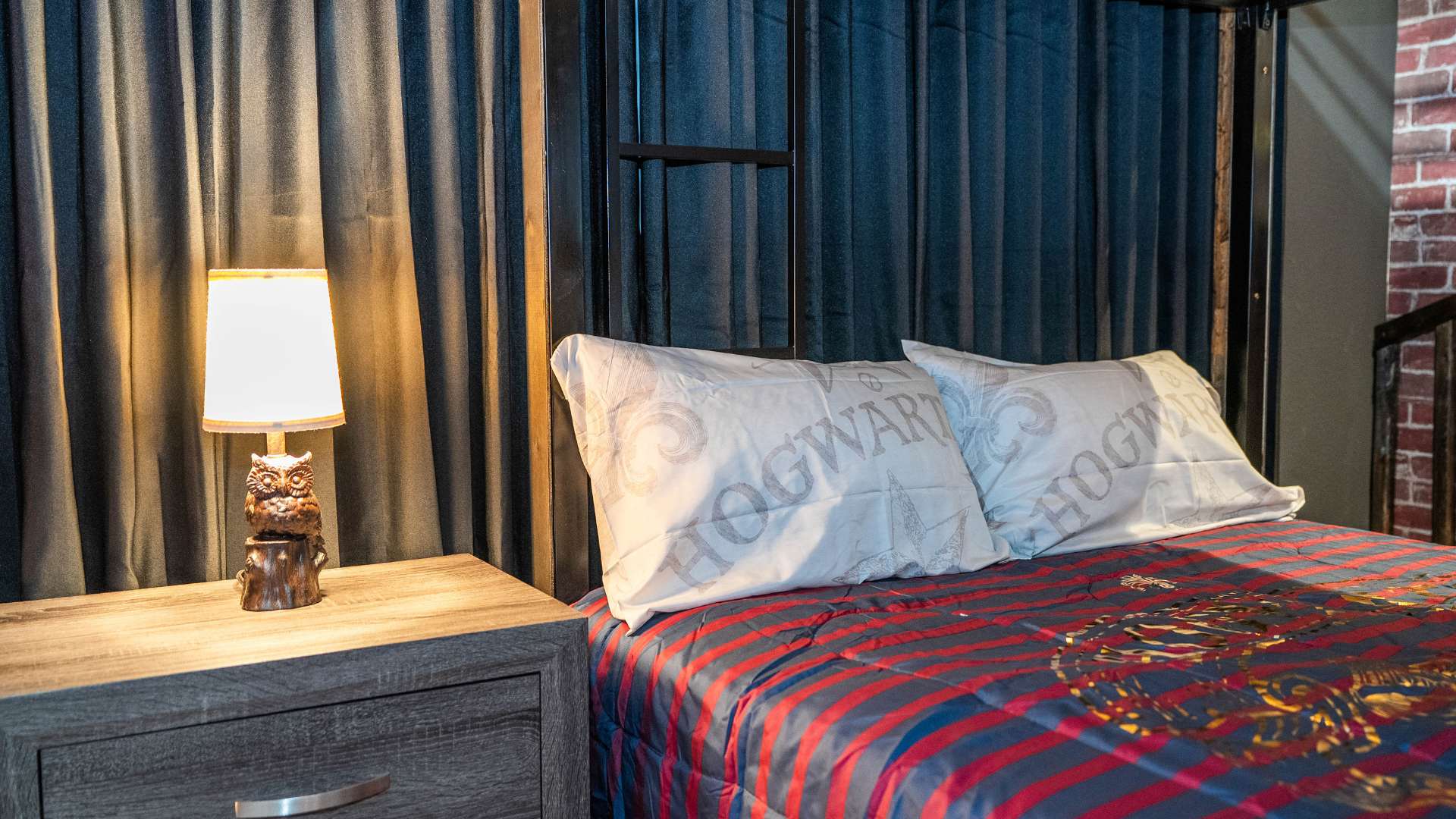 Lower bunk of the Harry Potter themed bedroom with striped bedding, "Hogwarts" pillows, an owl lamp on a nightstand, and dark blue pleated curtains.