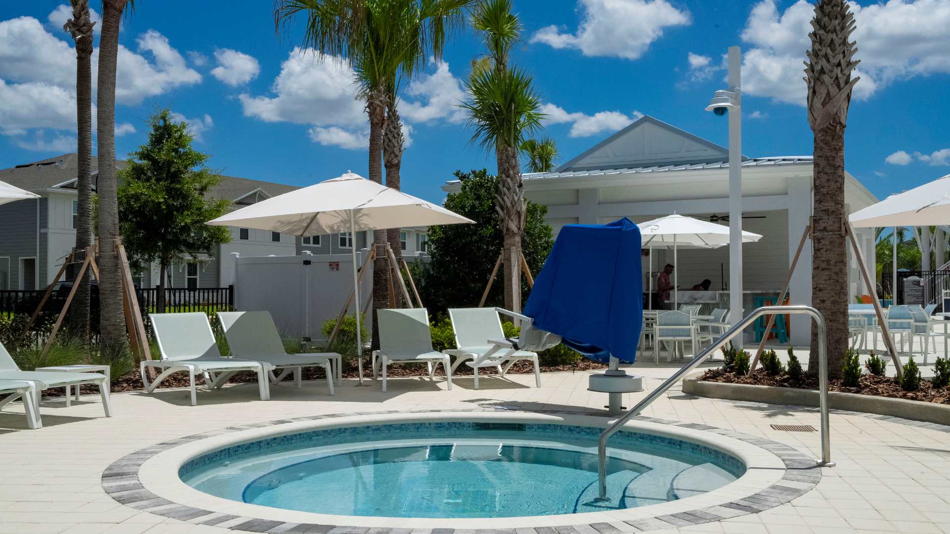 A circular hot tub with an accessible blue lift, surrounded by white lounge chairs and umbrellas on a paved patio, with resort buildings visible in the background under a sunny sky.