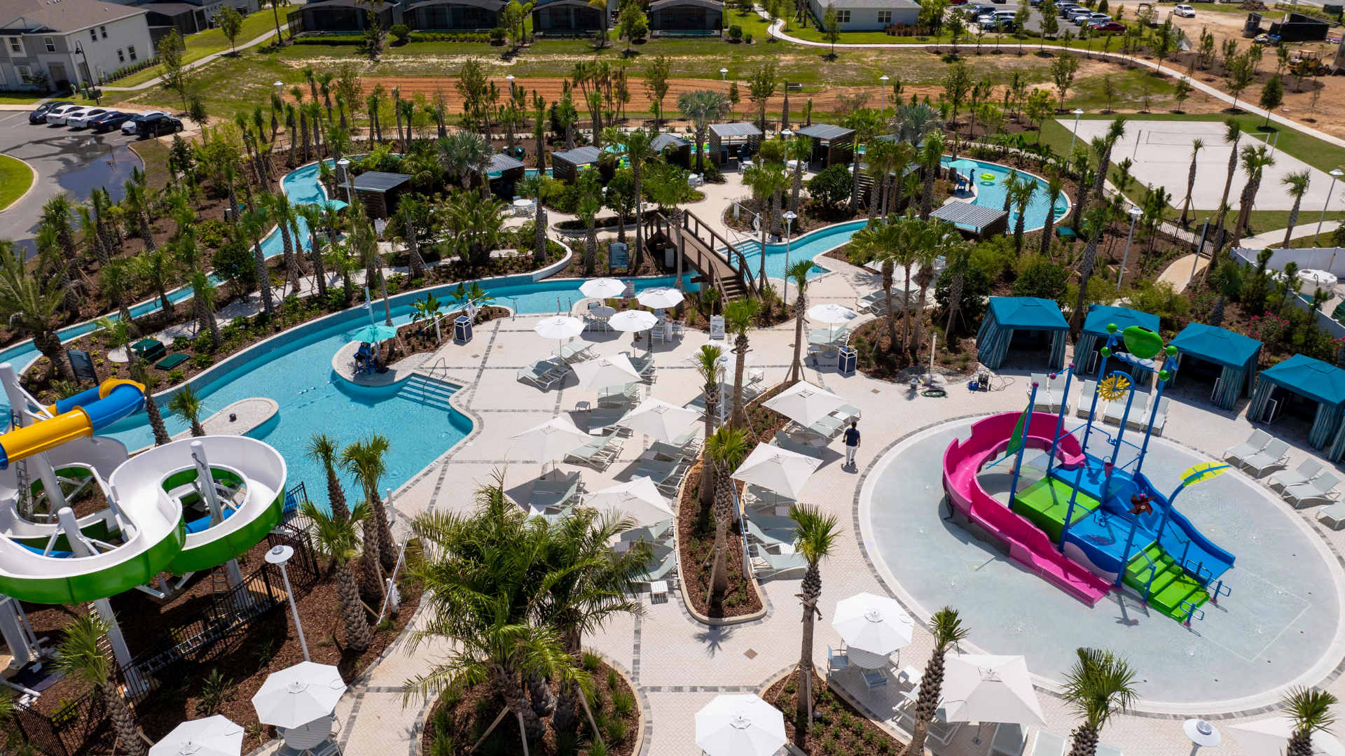 Expansive aerial view of a vibrant communal pool complex featuring a large swimming pool, a lazy river, multiple colorful water slides, numerous lounge chairs with umbrellas, and lush tropical landscaping.