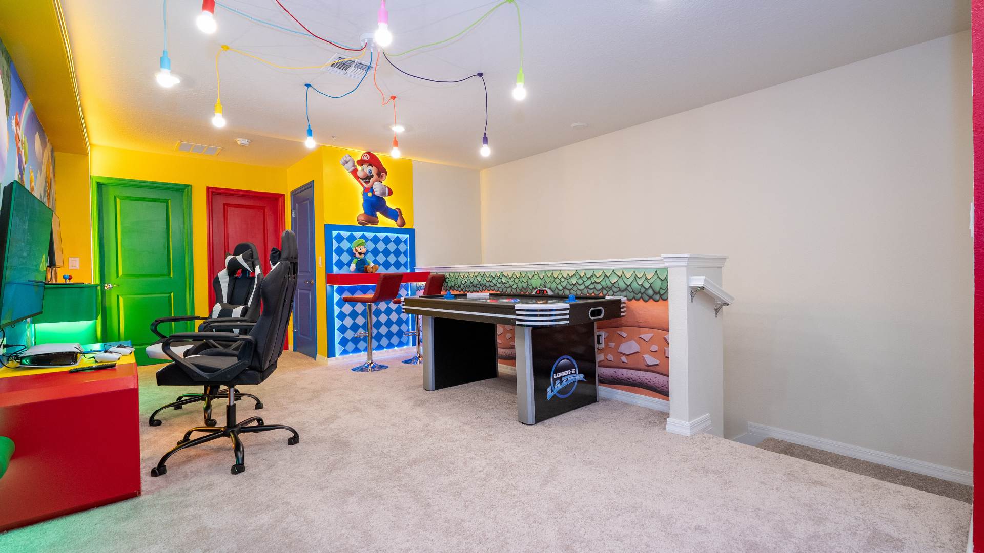 A spacious game room with light carpet features an air hockey table, two black gaming chairs, and a vibrant Mario wall mural. Colorful doors and a unique ceiling light fixture with exposed bulbs add to the playful and inviting atmosphere.