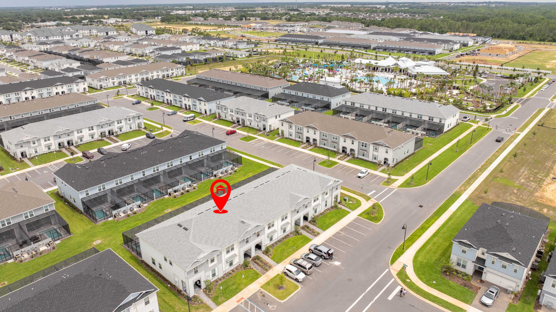 An aerial view captures a modern townhouse community with numerous light-colored buildings and green spaces. A red pin highlights the specific property, which is part of a row of townhouses, with a large communal pool area visible in the distance.