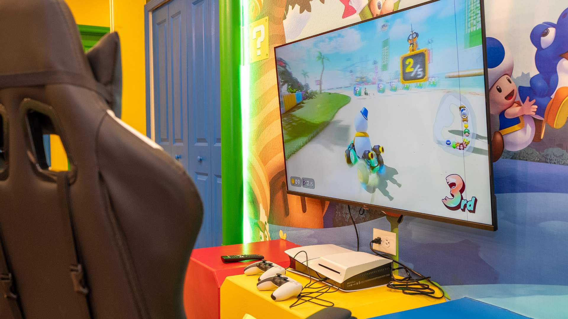 Close-up of a large TV displaying a Mario Kart game, with a white game console and two white controllers on a red and yellow block-themed stand. A black gaming chair is partially visible in the foreground, complementing the vibrant Mario-themed wall mural.