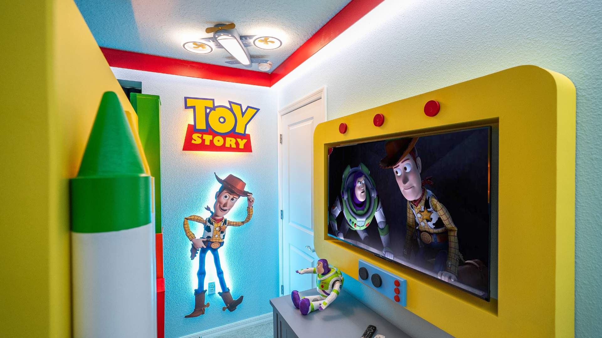 Immersive Toy Story themed bedroom with a wall-mounted TV framed by a yellow block design, a Woody cutout, and a Buzz Lightyear figure, all under a playful ceiling light.
