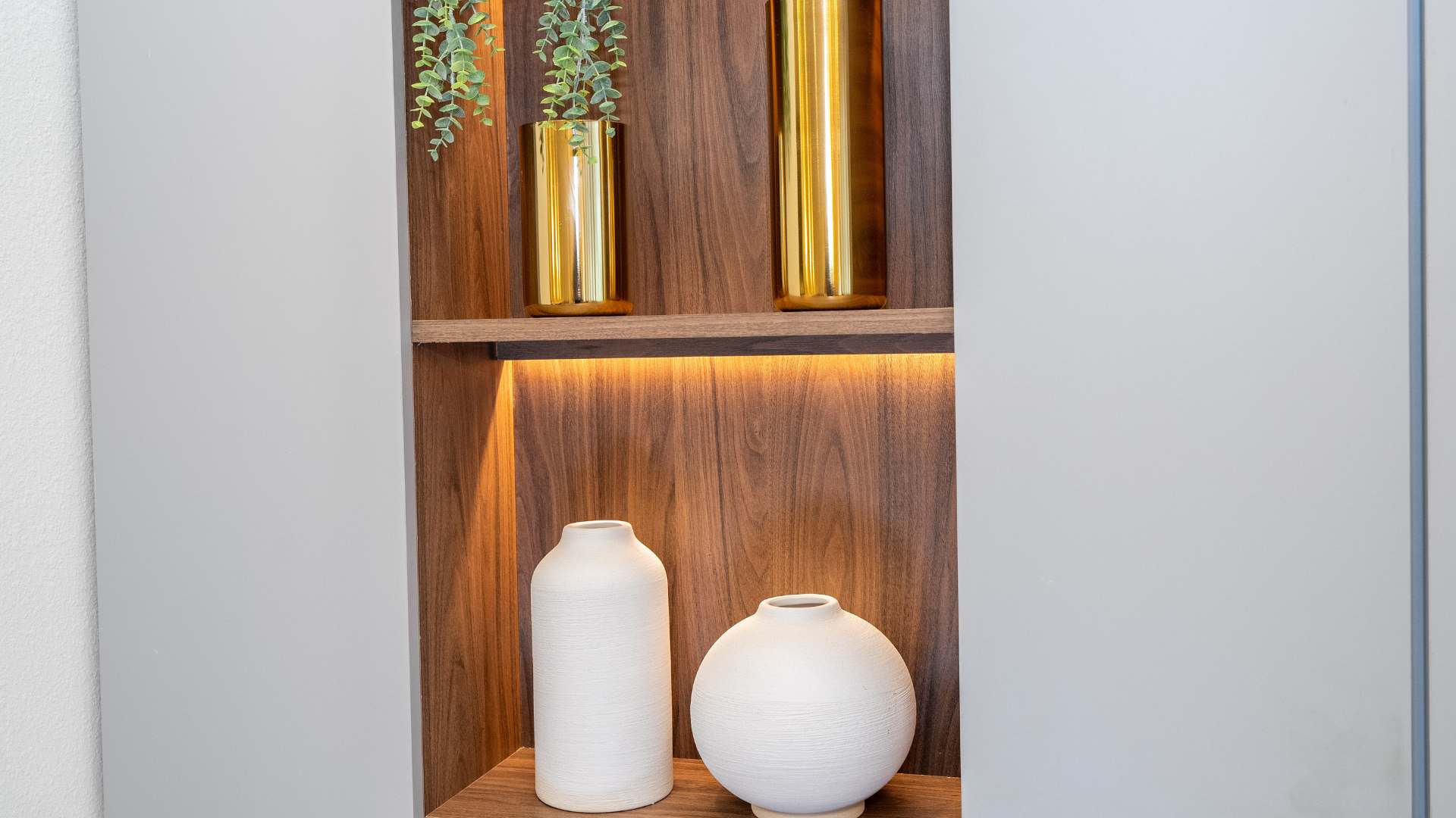 A decorative wall niche features two wooden shelves with integrated warm lighting, showcasing various decor items. The top shelf holds two tall gold vases and artificial greenery, while the bottom shelf displays two white textured ceramic vases.
