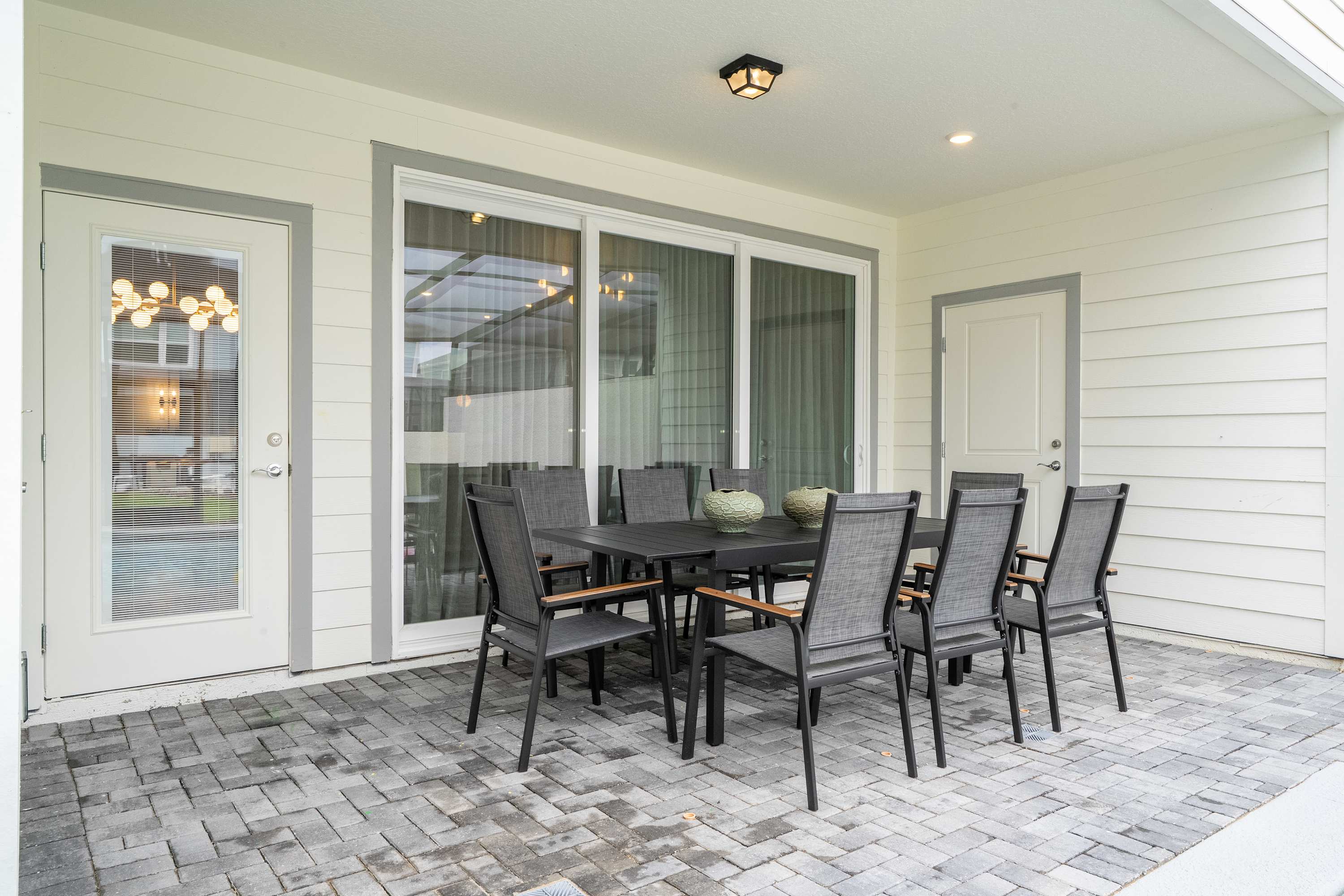 A spacious covered patio features a dark gray outdoor dining set with seating for six, positioned against the house with large sliding glass doors leading indoors. The area is paved with gray bricks and includes a white exterior door.