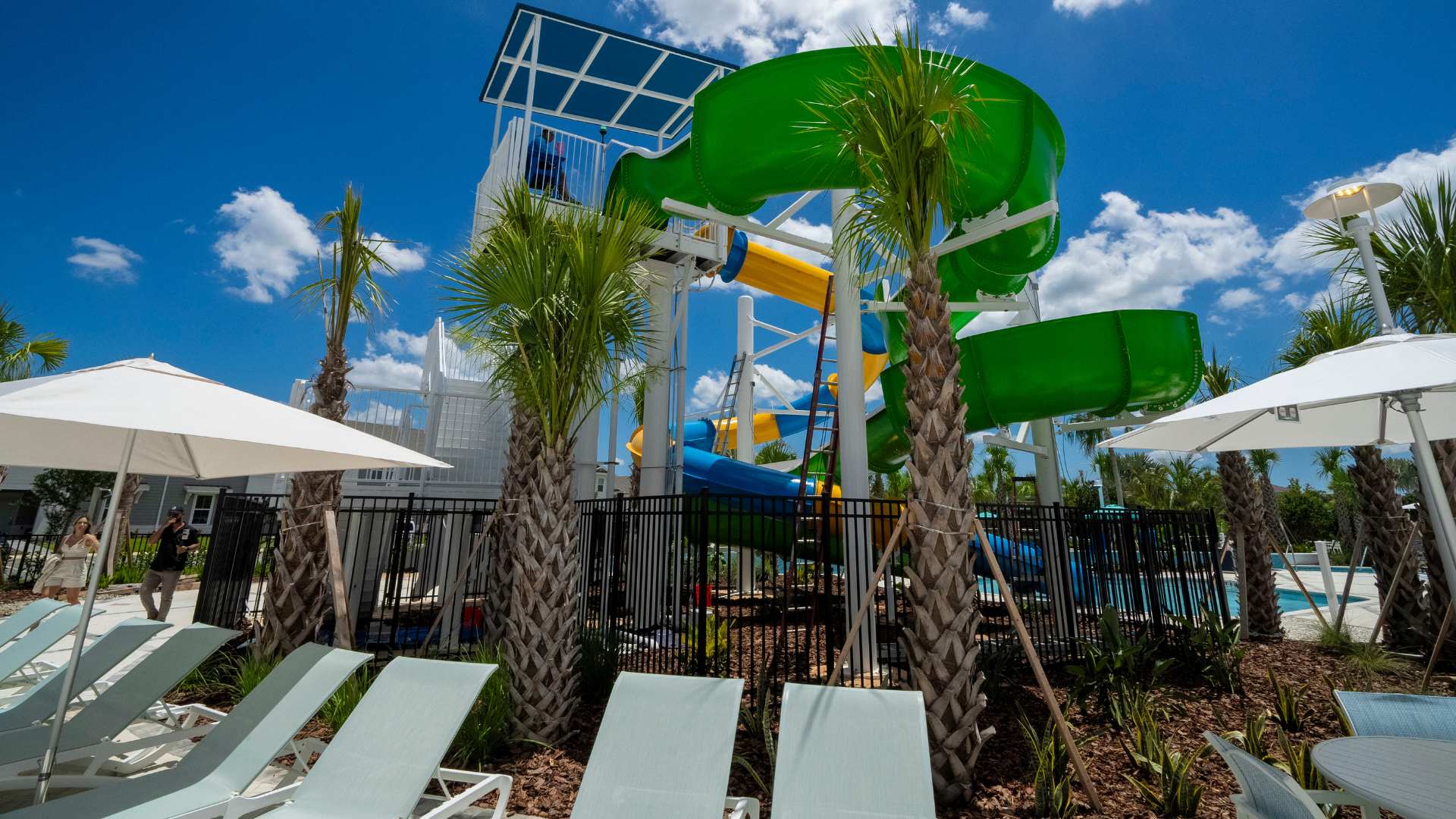 A detailed view of the multi-story water slide complex with green, yellow, and blue slides, featuring a white staircase and surrounded by tall palm trees, set against a clear blue sky.