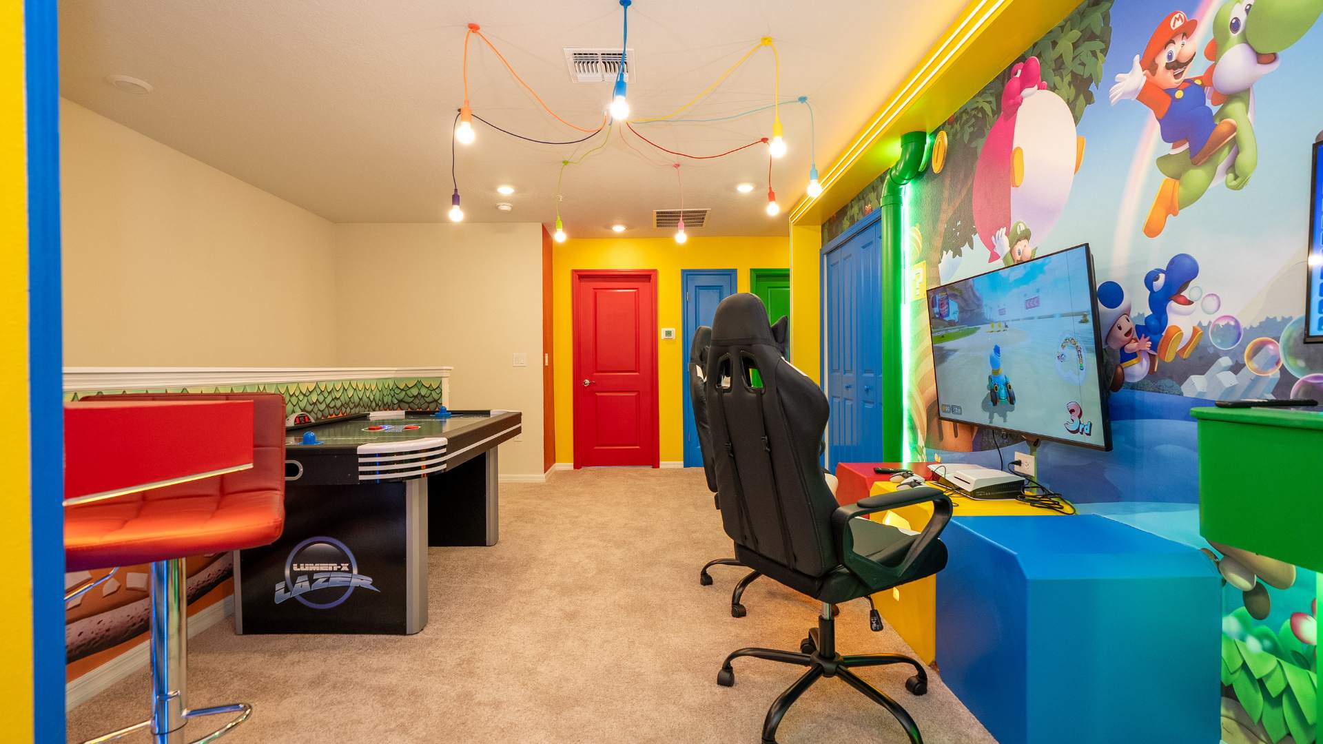 A vibrant game room with Mario-themed wall murals features a large TV displaying a video game, two black gaming chairs, and a colorful light fixture with exposed bulbs. Bright red, blue, and green doors add to the playful atmosphere.