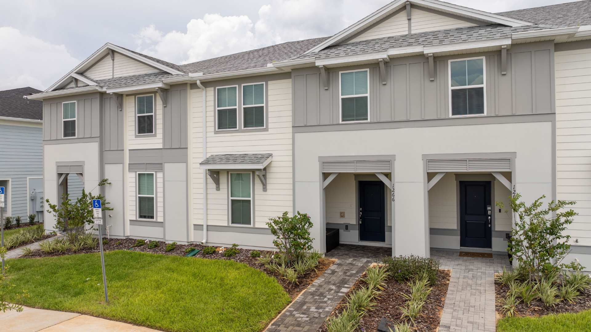 The front exterior of a modern townhouse features light-colored siding with gray accents and multiple windows. A paved walkway leads to the dark blue front door, surrounded by low-maintenance landscaping and a small covered porch area.