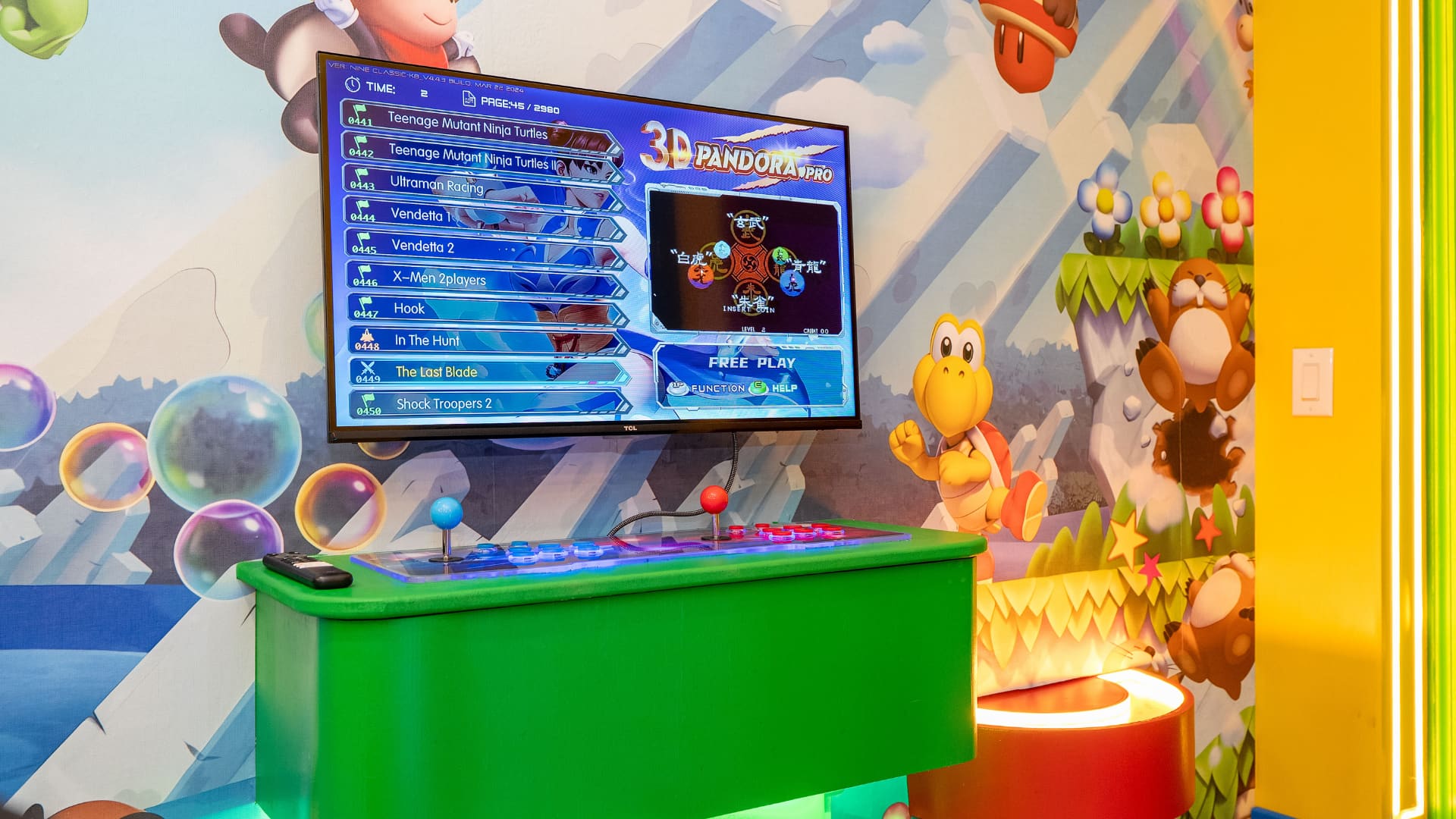 A close-up of the game room shows a wall-mounted TV displaying an arcade game menu, positioned above a green arcade cabinet with joysticks and buttons. The wall is adorned with a colorful Mario-themed mural featuring various characters.