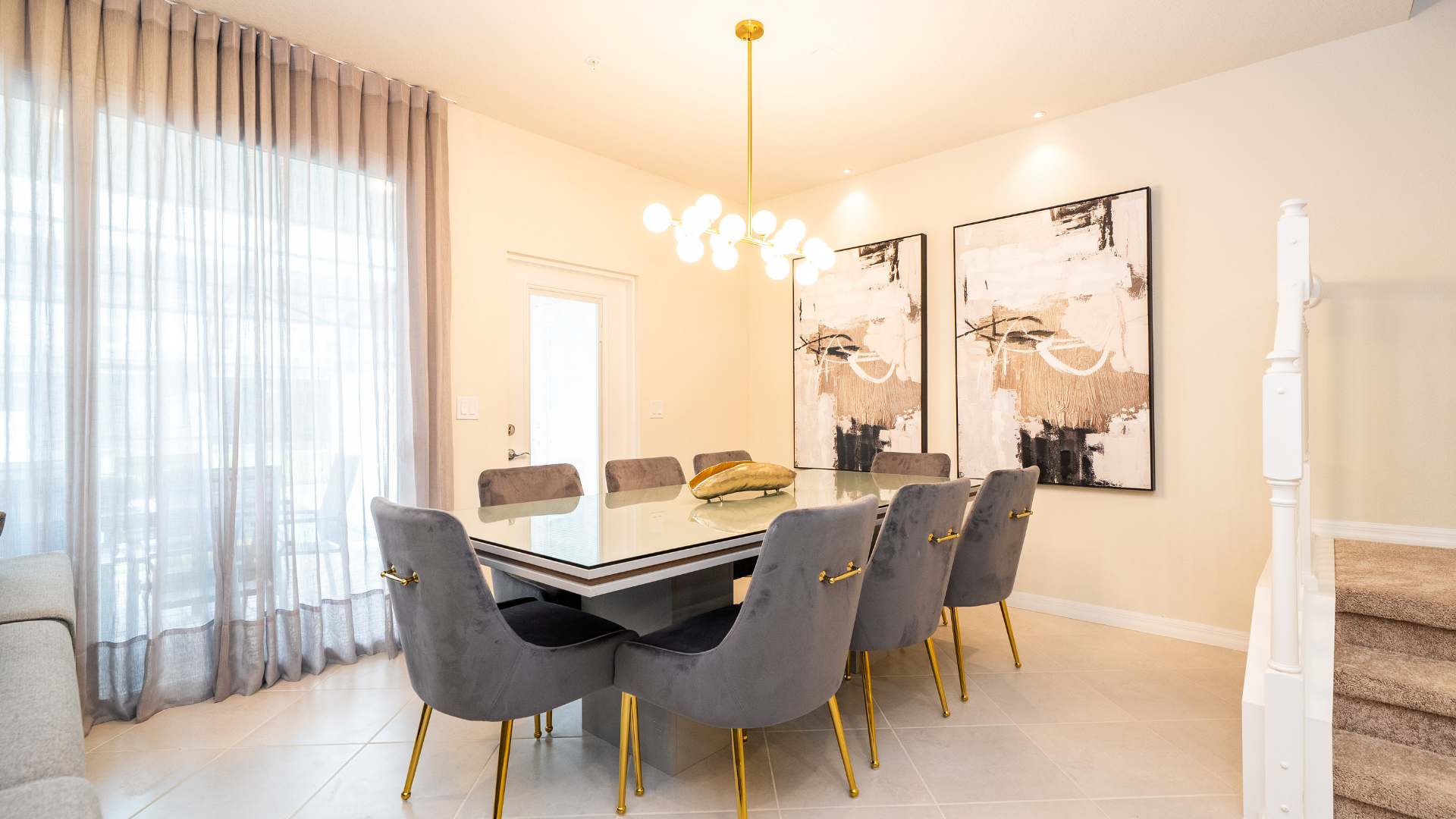 A modern dining room features a rectangular glass-top dining table with a gray base, surrounded by six gray velvet chairs with gold legs. Contemporary abstract artwork adorns the wall, and a gold multi-bulb chandelier hangs above the table.