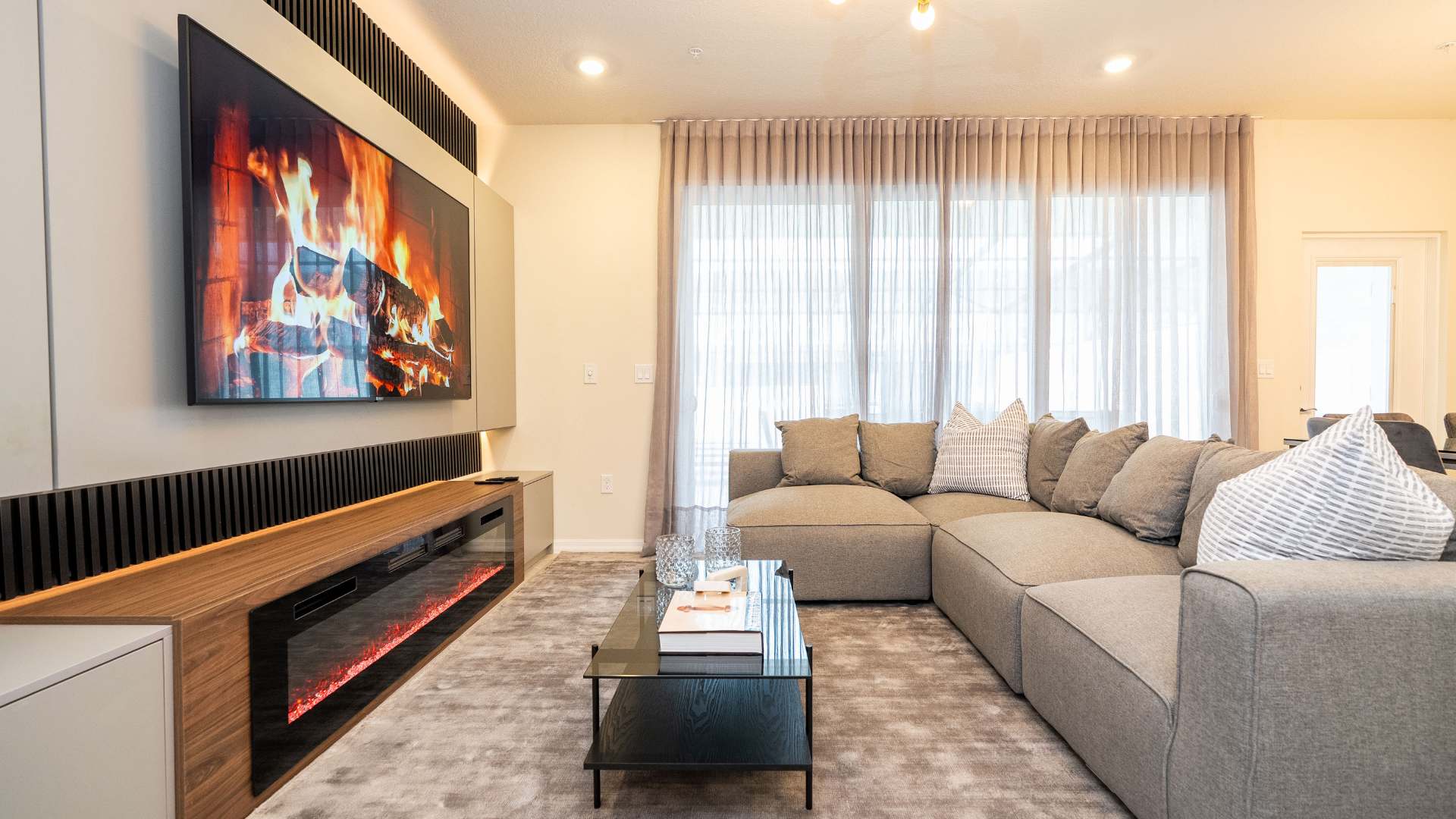 View of the modern living room featuring a large grey sectional sofa, a black glass coffee table, and a wall-mounted TV above an electric fireplace. Large windows with sheer curtains provide natural light, and a gold chandelier hangs from the ceiling.