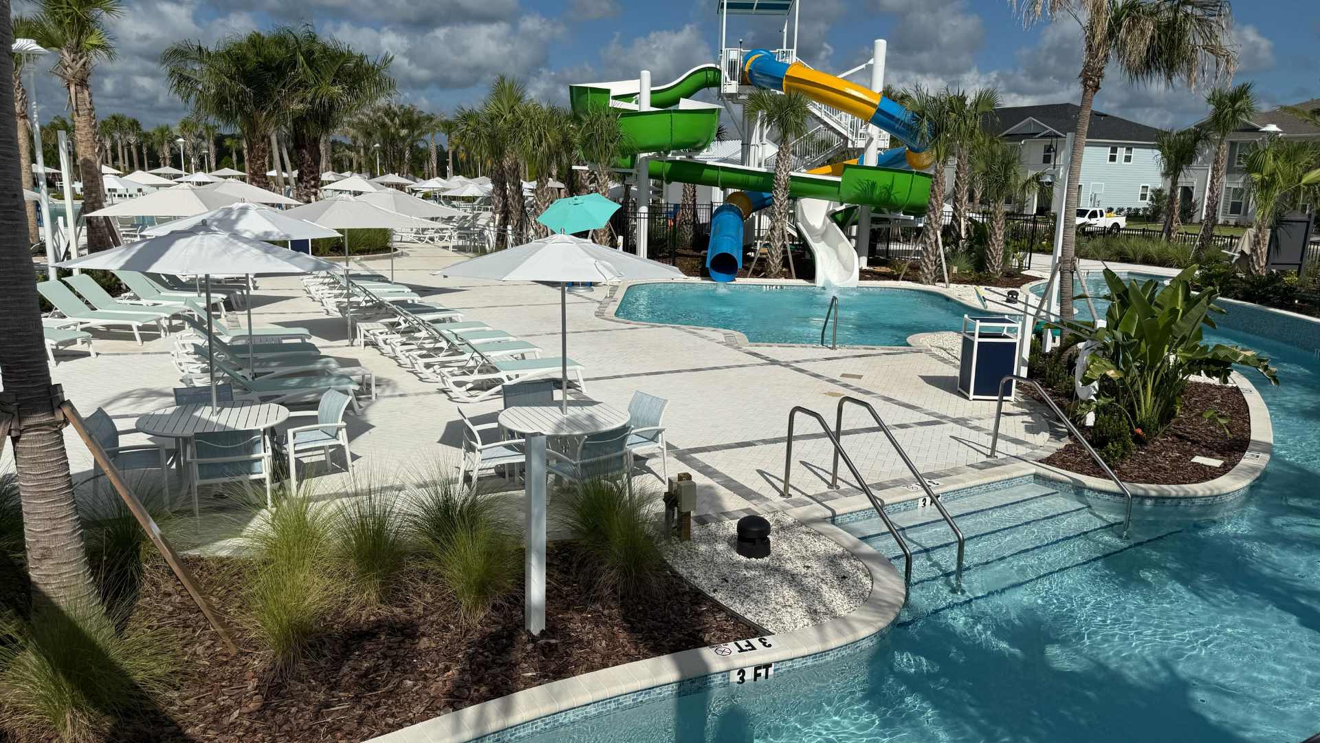 Vibrant communal pool area with a multi-colored water slide structure, a winding lazy river, and a spacious white paved deck furnished with light blue lounge chairs and white umbrellas.