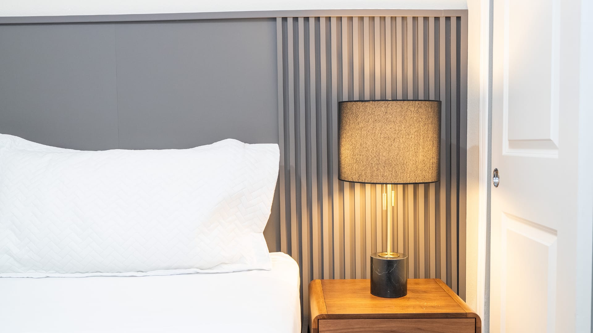 A close-up view of a modern bedside setup shows a dark grey textured lamp with a gold base on a wooden nightstand. White pillows and bedding are visible on the bed, next to a white bi-fold closet door.