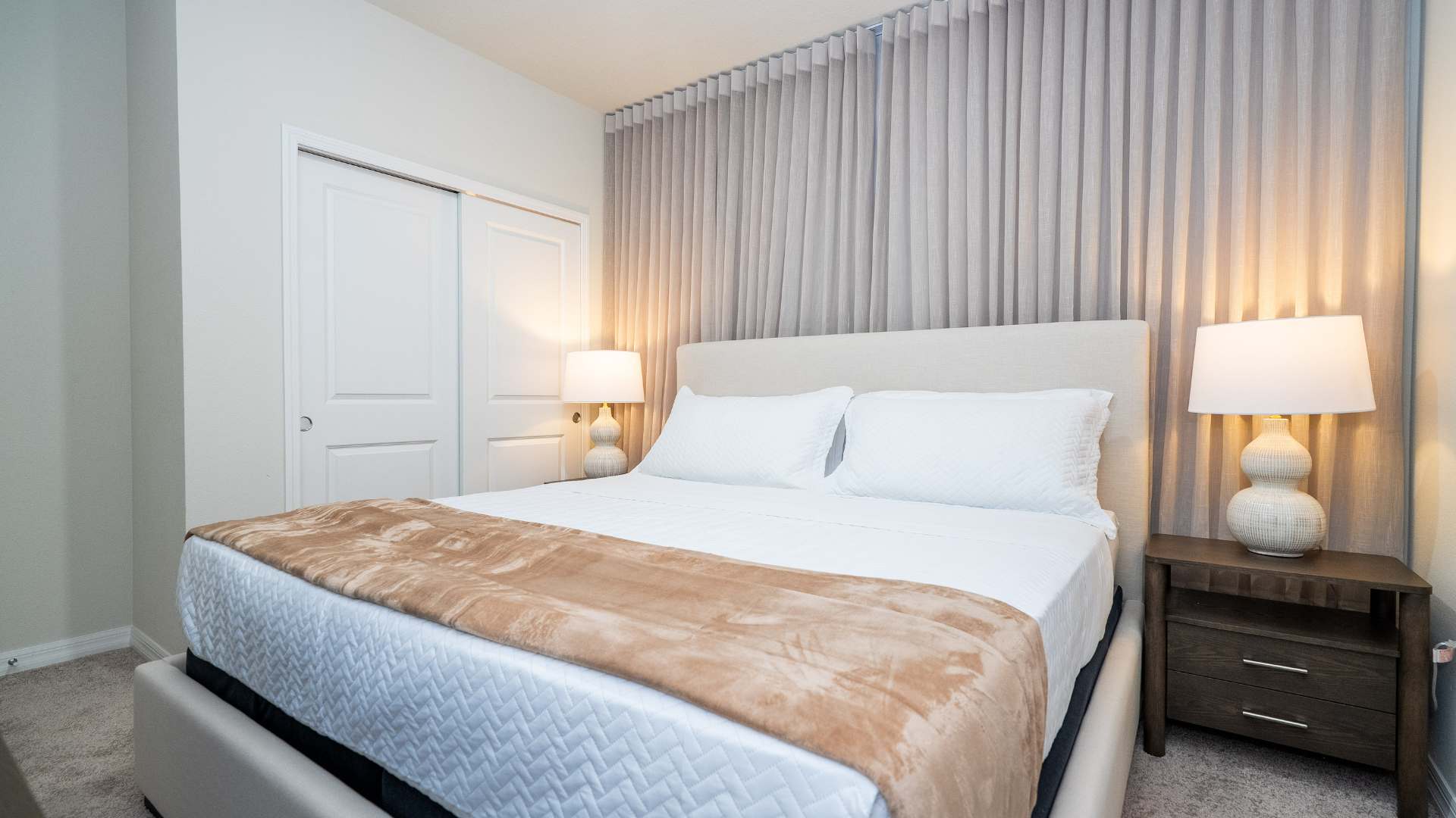 Cozy bedroom with a light beige upholstered bed, two nightstands with lamps, and a closet with bi-fold doors, all set against a backdrop of light gray vertical blinds.