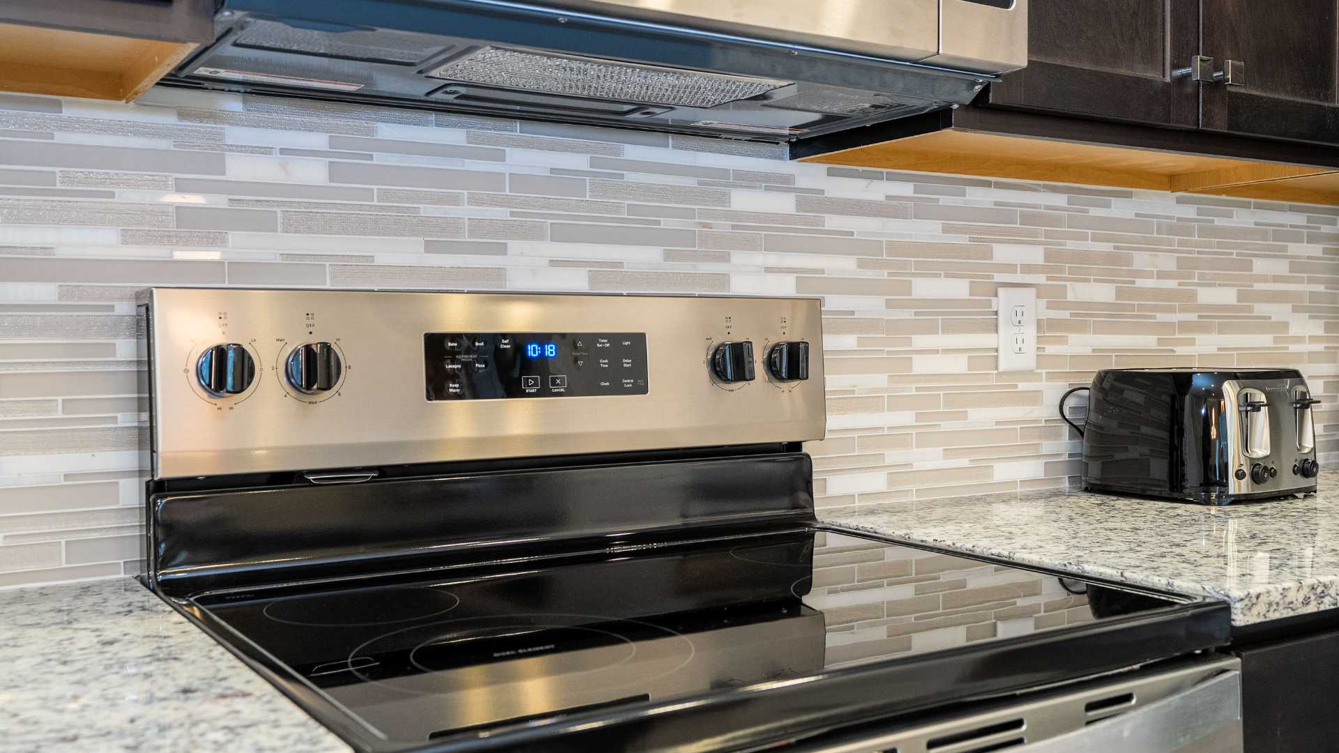 Close-up of a stainless steel electric range with a black cooktop and control panel, set against a light-toned mosaic tile backsplash. A toaster is visible on the granite countertop.