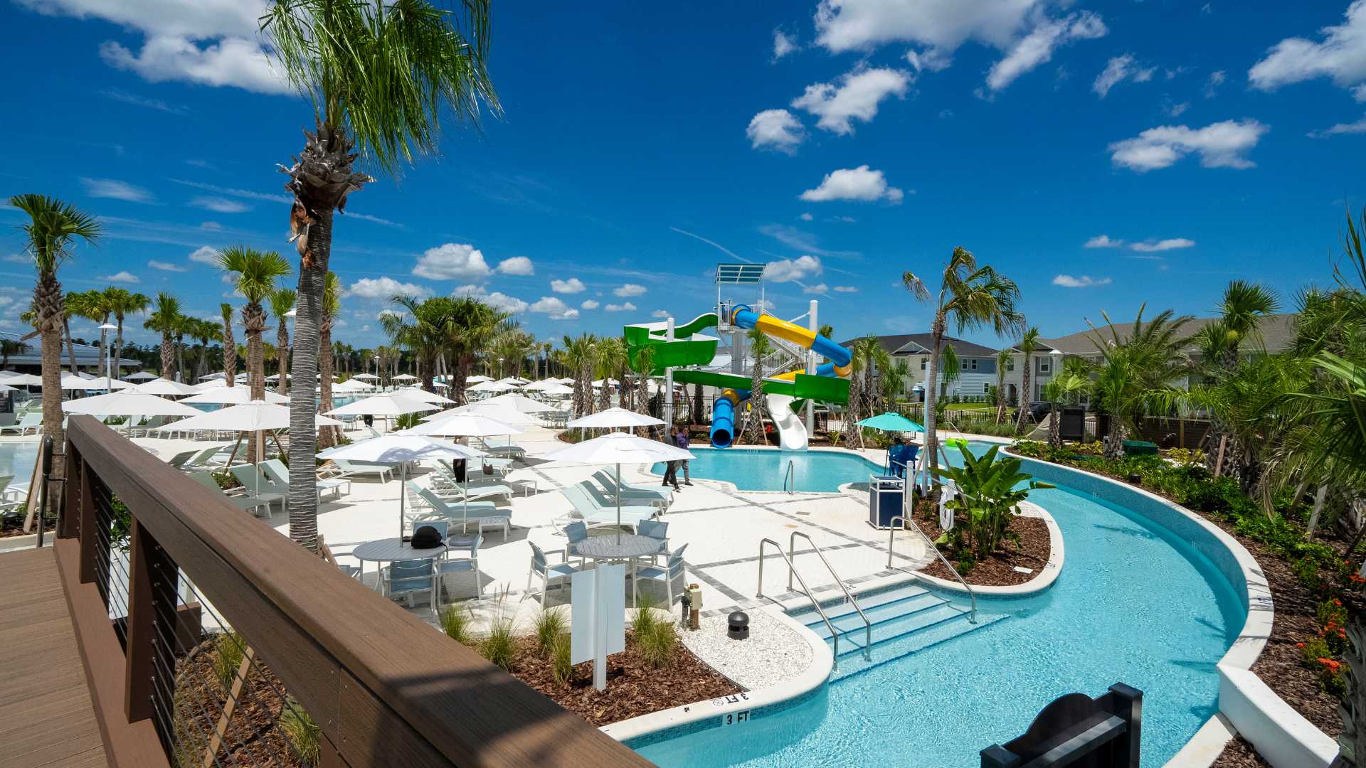 Expansive resort-style pool area featuring a winding lazy river, colorful water slides, and a large deck with numerous white lounge chairs and umbrellas under a bright blue sky.