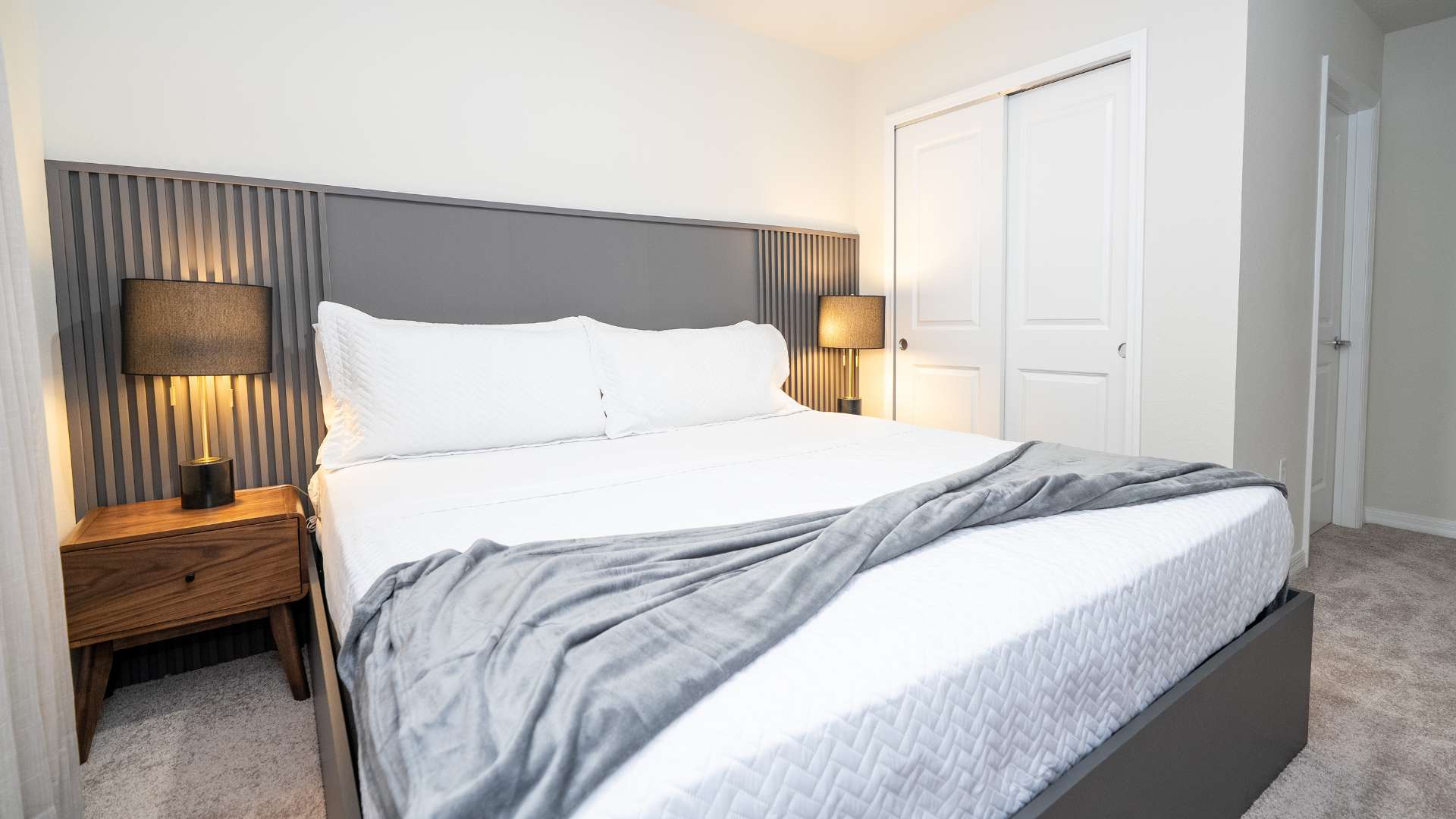 A modern bedroom features a large bed with white bedding and a grey throw, flanked by two wooden nightstands with black lamps. A grey slatted headboard extends across the wall, and a white bi-fold closet door is visible on the right.