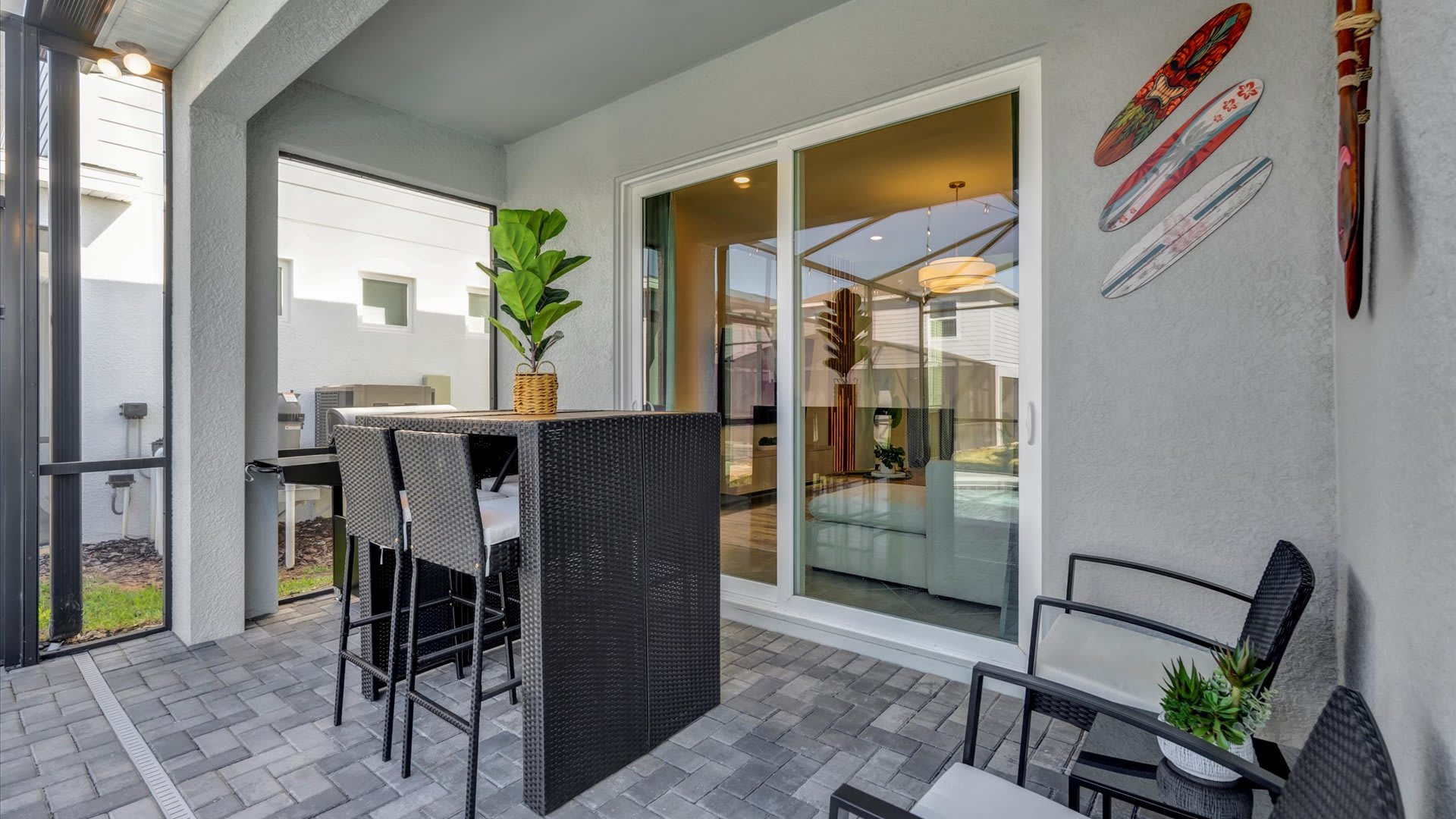 Covered outdoor patio area with grey paver flooring, featuring a tall black wicker bar table with stools and additional seating. Sliding glass doors lead into the interior living space, and decorative surfboards hang on the wall.