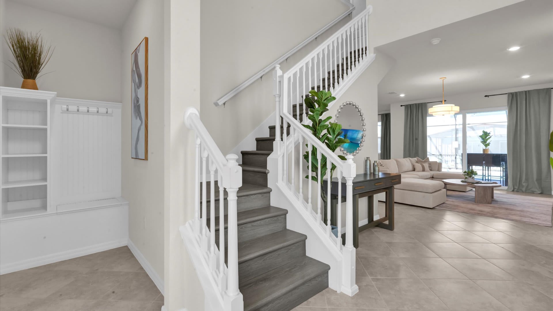 Grand foyer featuring a light gray wooden staircase with white railings, leading upstairs. A console table with a large round mirror and a plant sits beneath the stairs, with light tile flooring extending throughout the space.