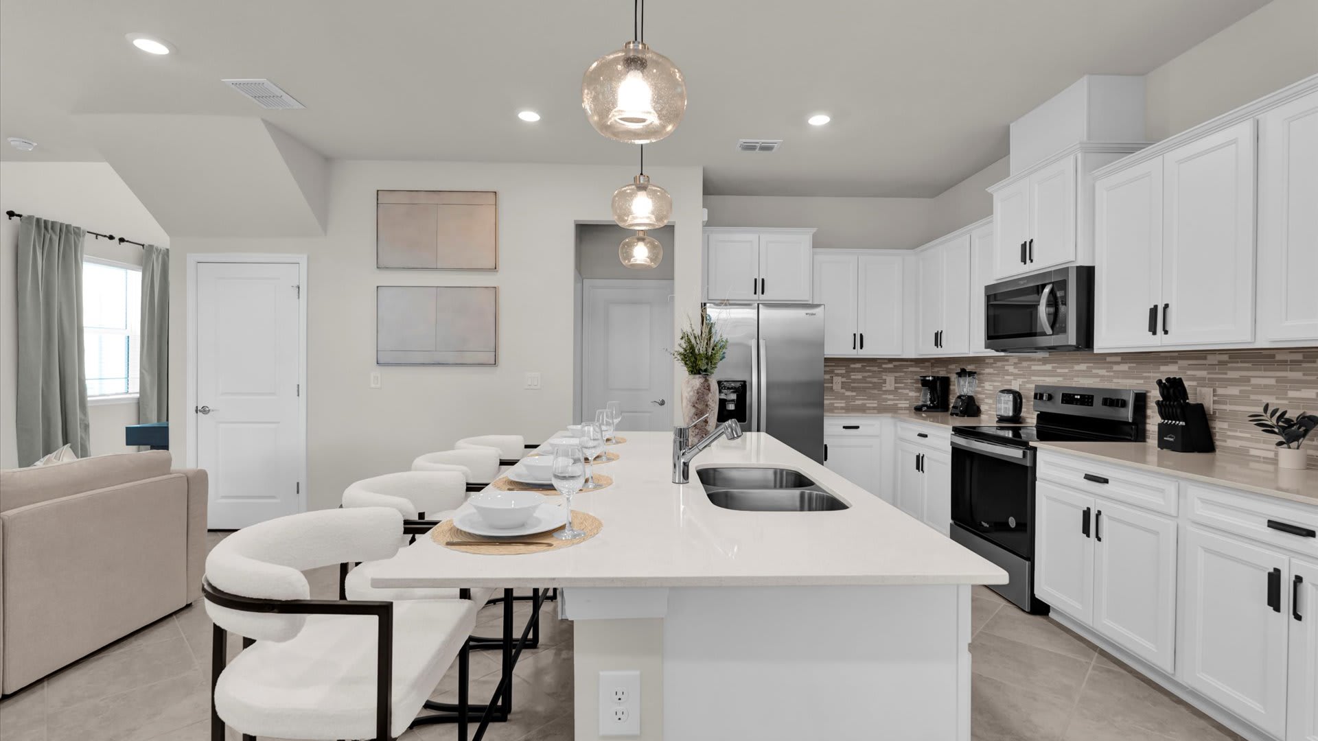 Bright kitchen island with a white countertop and four bar stools, offering a view into the adjacent living area. The kitchen features white cabinetry, stainless steel appliances, and light tile flooring, maintaining a cohesive modern aesthetic.