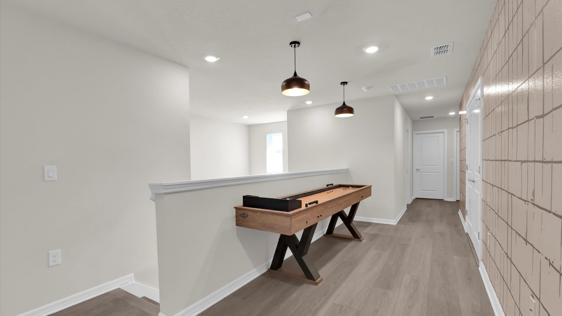 Upstairs landing converted into a game area, featuring a wooden shuffleboard table with black accents. The space has light wood-look flooring, white walls, and modern pendant lights, with a textured accent wall on the right.