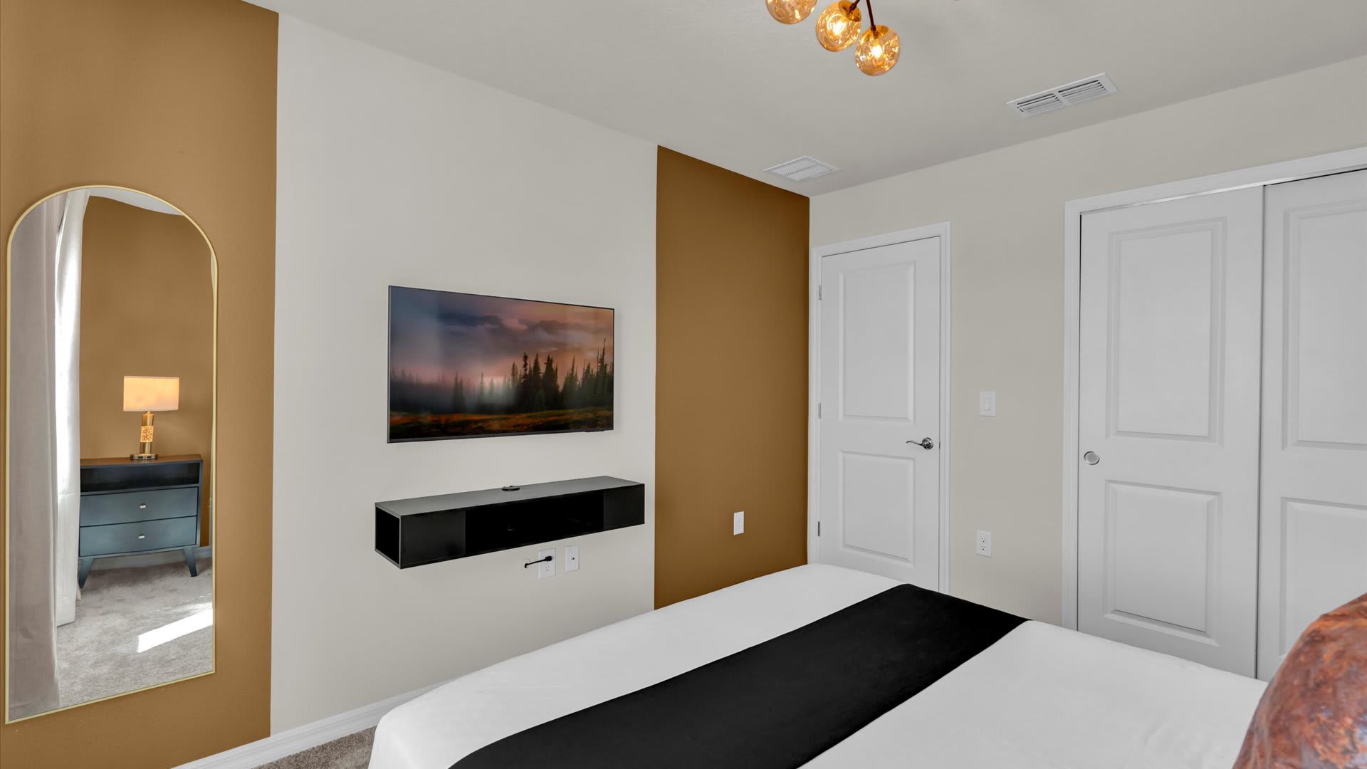 Another view of the bedroom, showing a wall-mounted TV above a black floating shelf. A full-length arched mirror leans against the gold accent wall, and two white doors are visible.
