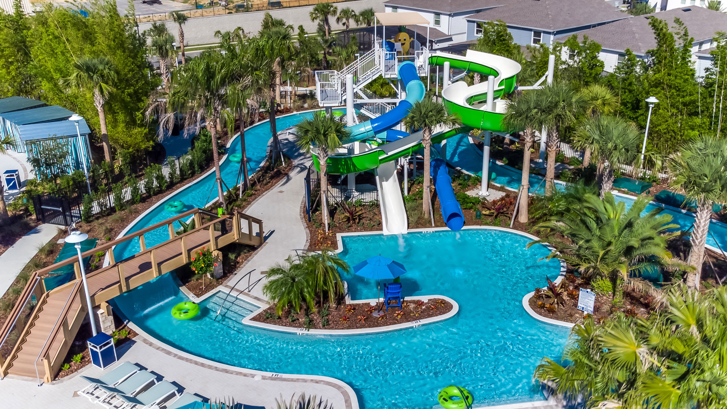 An aerial view of a vibrant communal pool area featuring a lazy river, multiple water slides in blue and green, and lush palm trees. A wooden bridge crosses the lazy river, and lounge chairs are visible on the surrounding deck.