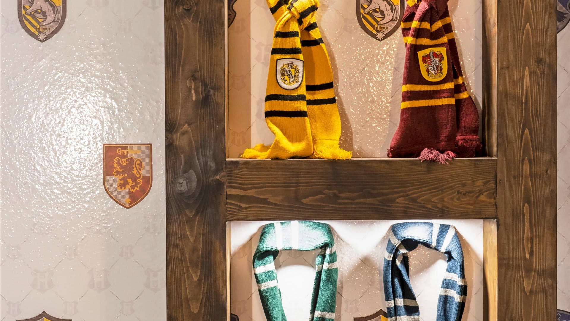A themed display wall featuring shelves with various colored scarves, including yellow and black, and maroon and gold, each with house crests. The wall is decorated with subtle crest patterns and wooden accents.