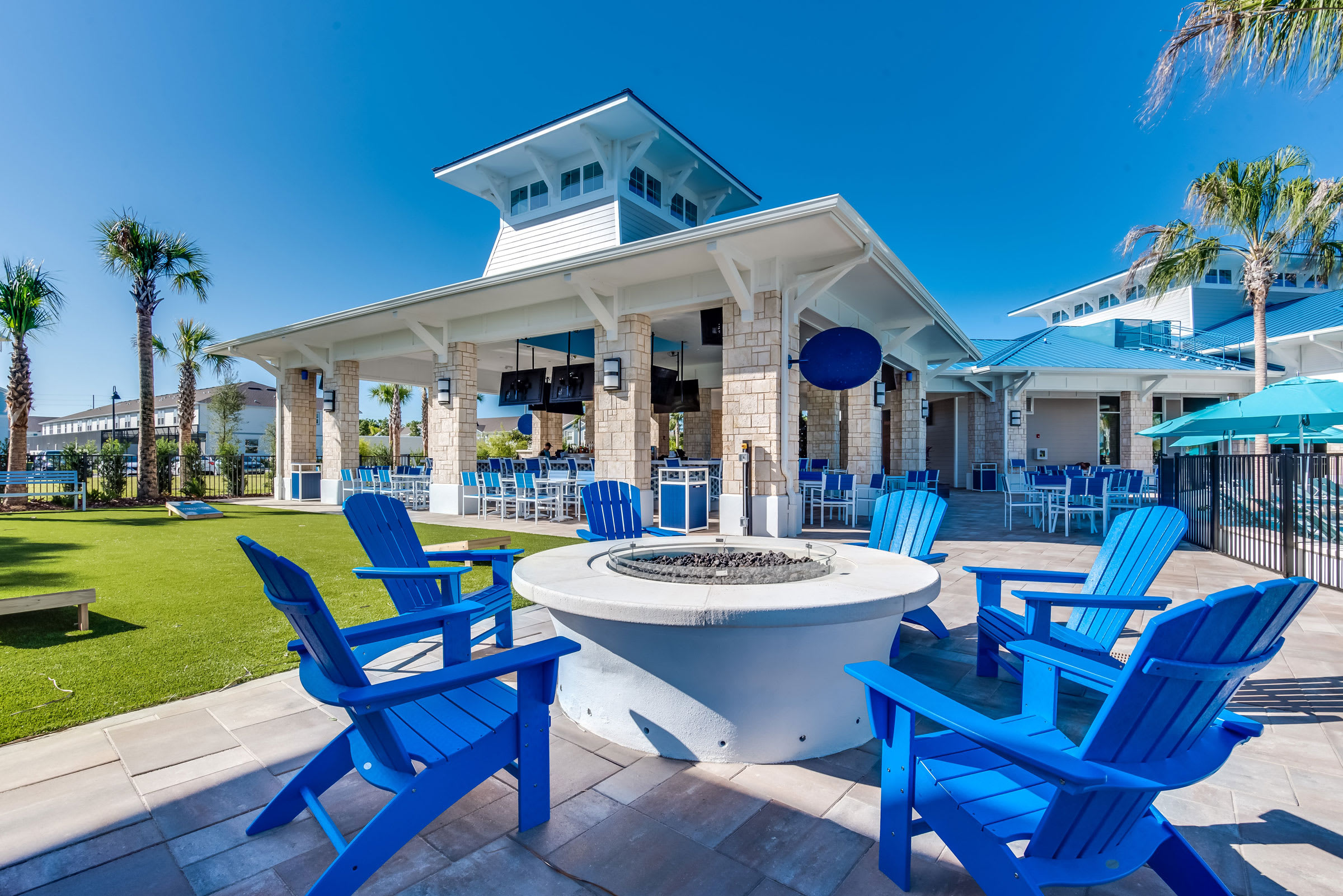 An inviting outdoor fire pit area features a circular white fire pit surrounded by bright blue Adirondack chairs, set on a paver patio adjacent to a lush green lawn. In the background, a large covered pavilion with a bar and additional seating offers a communal gathering space under a clear blue sky.