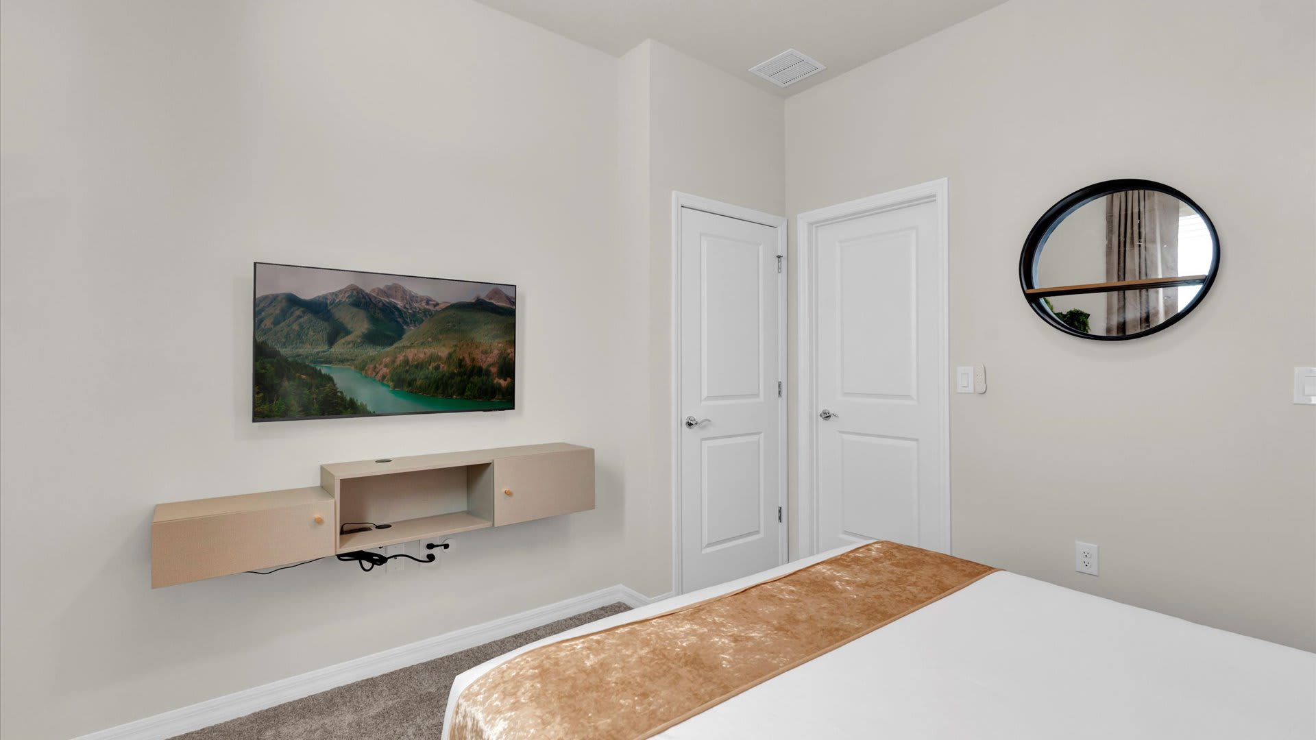 Another angle of the bedroom, showing a wall-mounted TV above a floating wooden console. A round black-framed mirror hangs on the adjacent wall, and two white doors are visible.