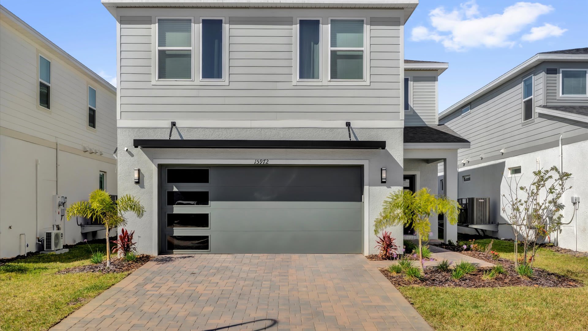 Modern two-story house with light grey siding and a dark grey garage door, featuring a paved driveway and small landscaped areas. The house number '15912' is visible above the garage.