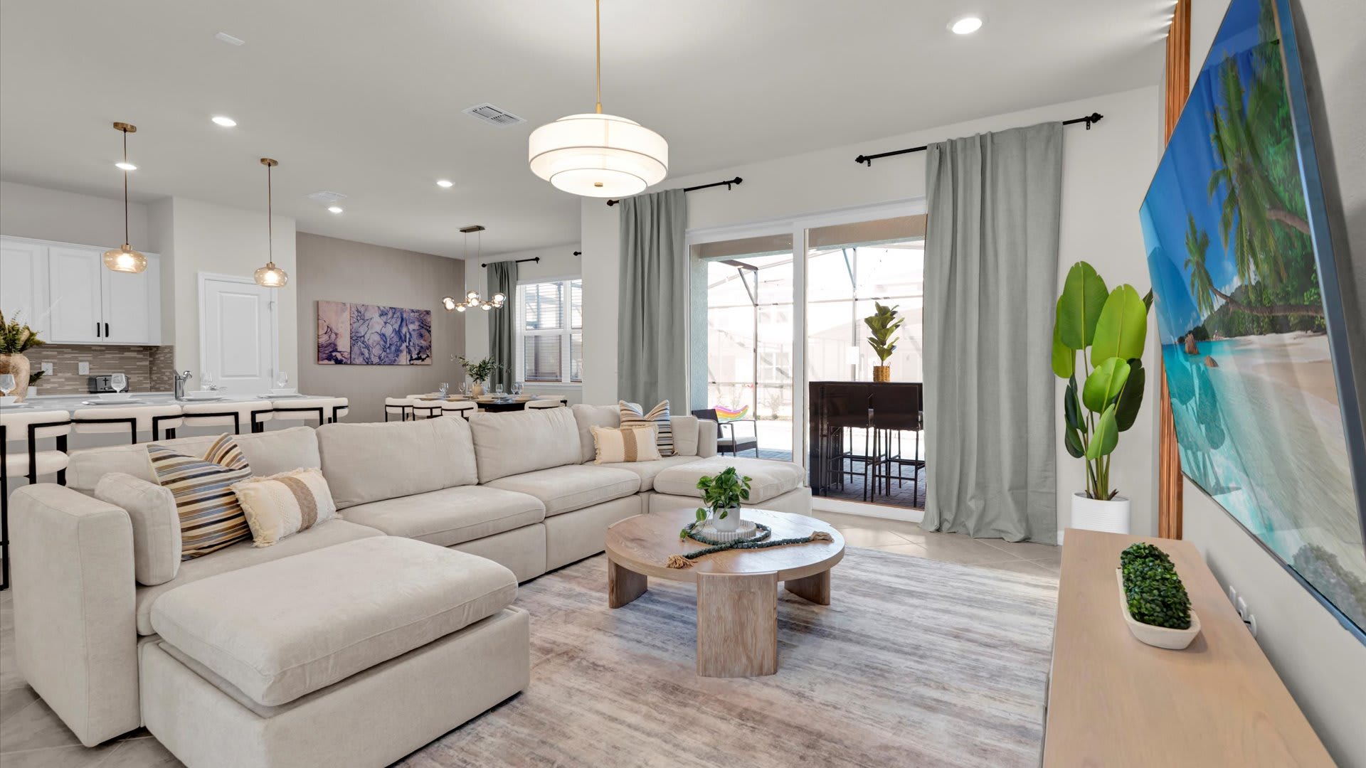 Bright and open-concept living room with a large L-shaped sectional sofa in a neutral tone, a round wooden coffee table, and a patterned area rug. Sliding glass doors lead to the outdoor patio and pool area, and a large TV is mounted on the wall.