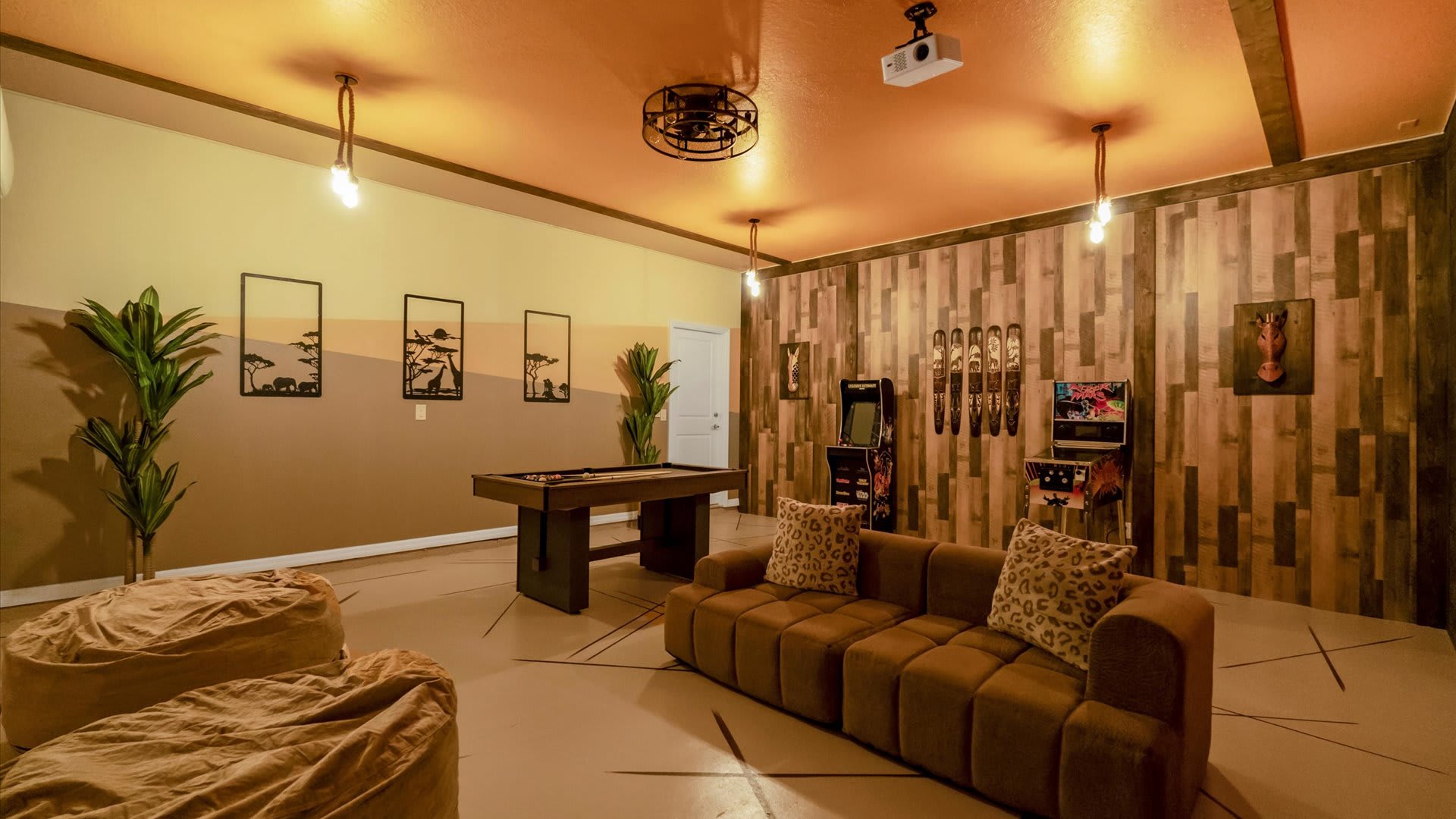 Wide view of the game room featuring a brown sofa with leopard print pillows, a pool table, and two bean bag chairs. The room has warm-toned walls, wood accents, and safari-themed decor, including metal wall art and a wooden horse head.