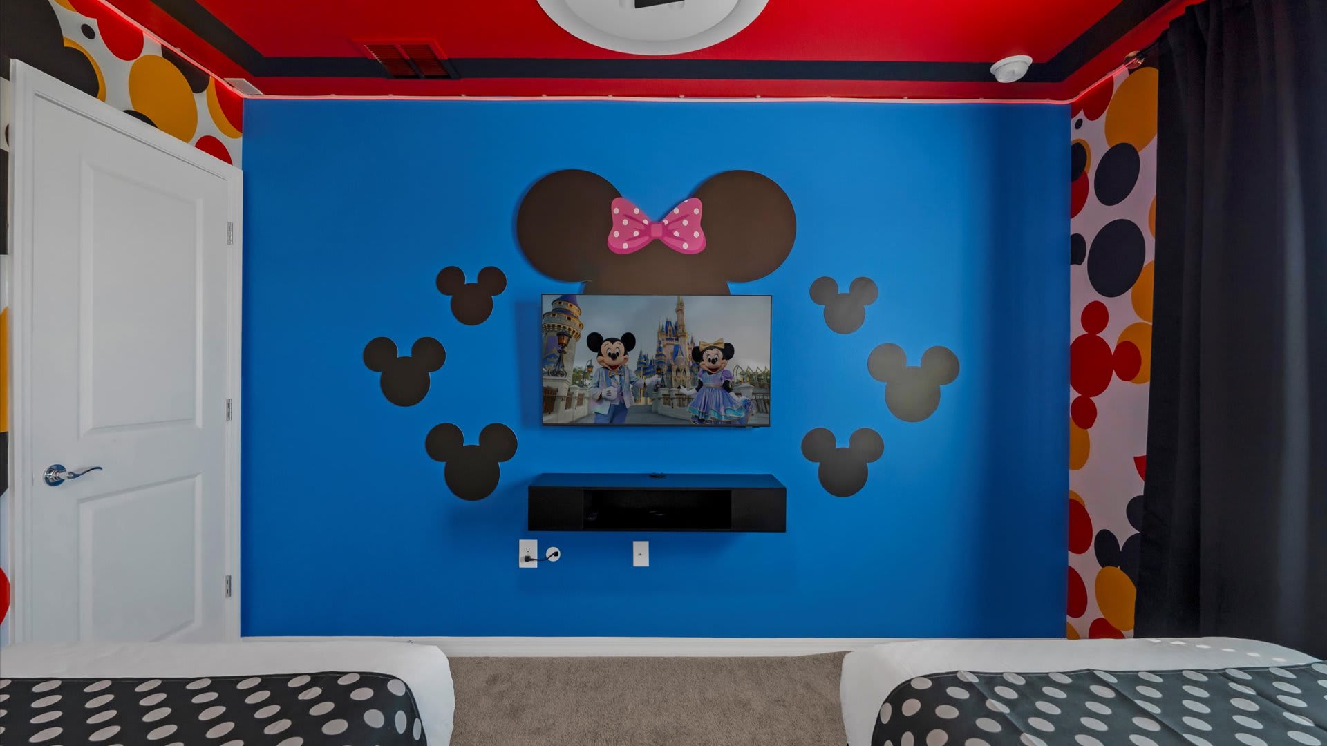 A vibrant blue themed bedroom featuring a large Minnie Mouse head with a pink bow above a wall-mounted TV, flanked by smaller Mickey Mouse head cutouts. Two beds with black and white polka dot bedding are partially visible in the foreground.