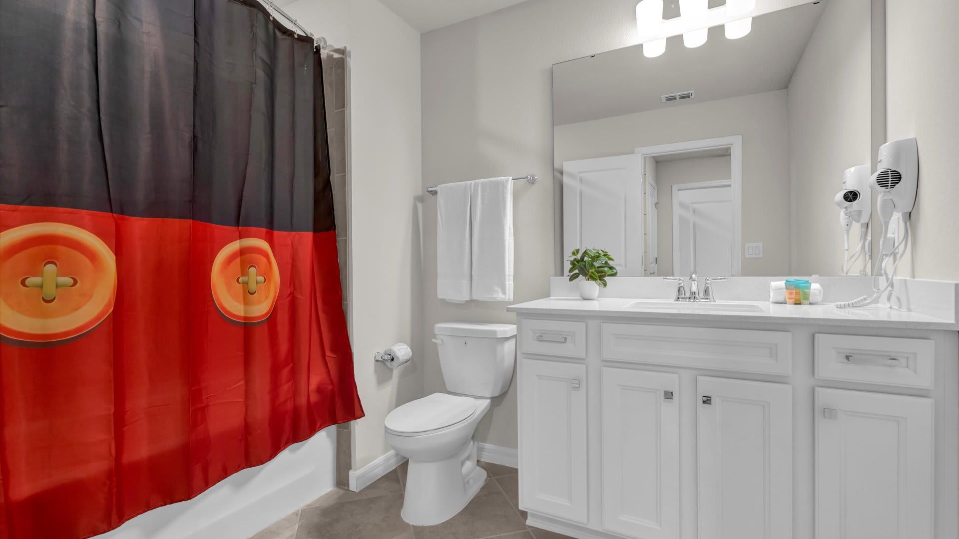 A bright bathroom with a white vanity, sink, and mirror, featuring a Mickey Mouse themed shower curtain in red and black with large yellow buttons. A toilet is visible next to the vanity.