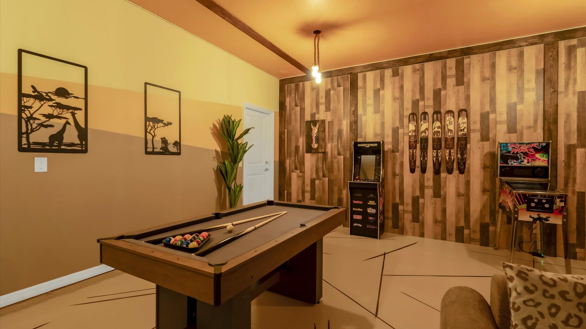 Themed game room with a pool table in the center, flanked by arcade machines and decorated with safari-themed wall art. One wall features vertical wood paneling, while another has a two-tone painted design.