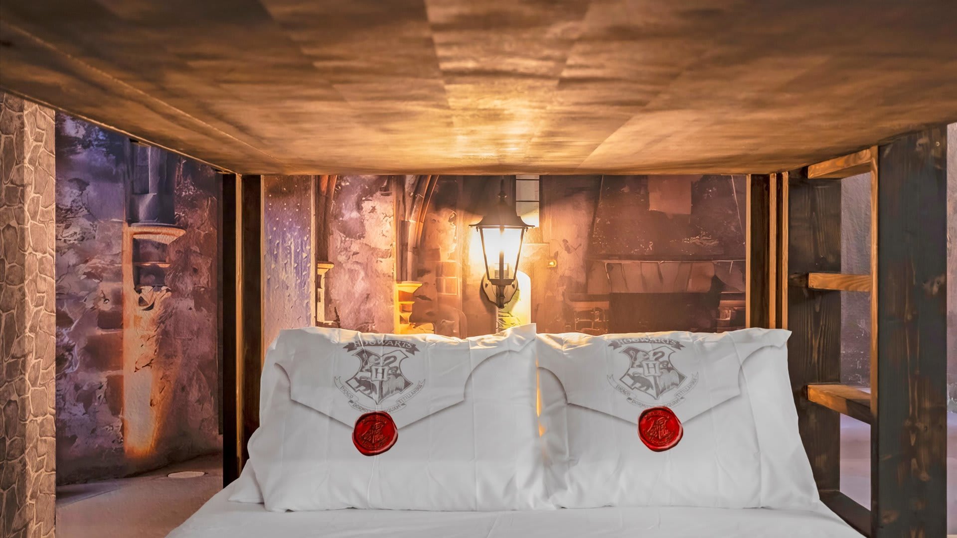 A close-up view of the lower bunk bed in a Harry Potter-themed room, showcasing white pillows with Hogwarts crests and red wax seal designs. The background features a detailed mural of a stone wall with a lantern.