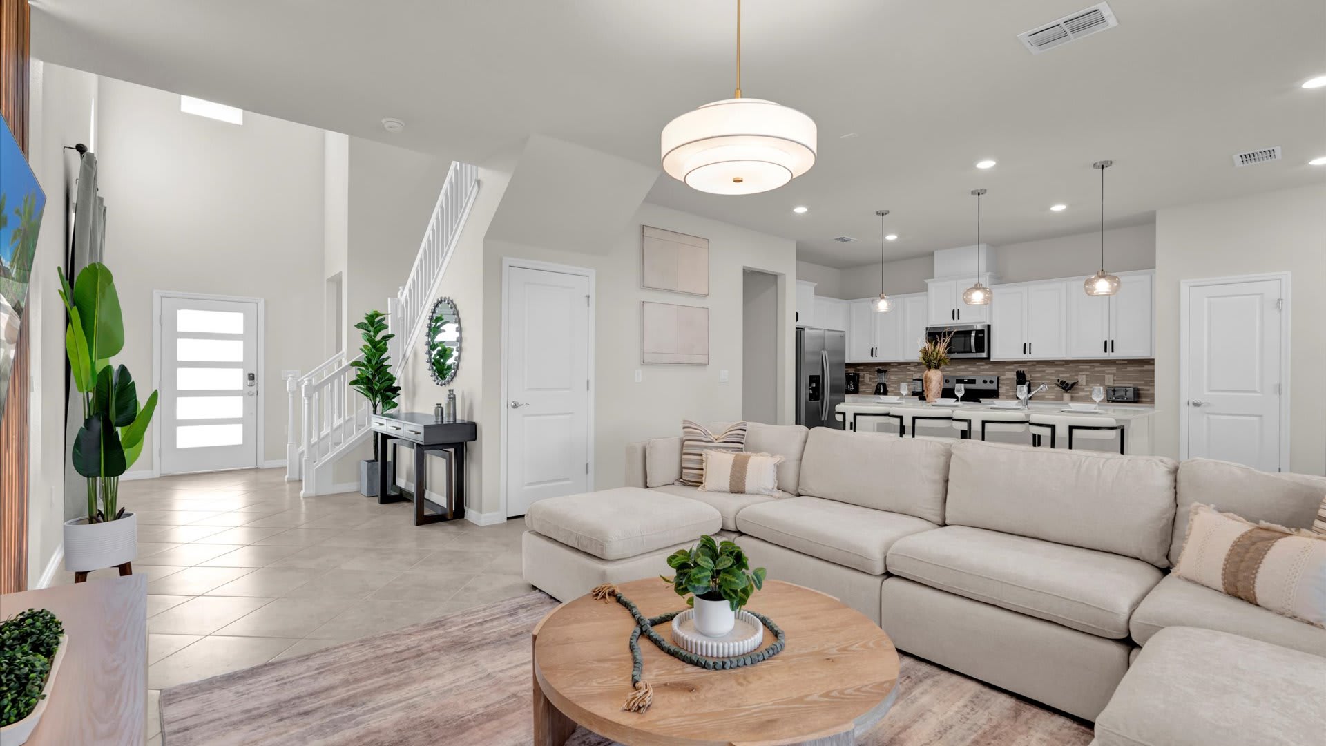 Bright, open-concept living area with a large L-shaped sectional sofa, a round wooden coffee table, and light tile flooring. The space opens to a modern kitchen and a staircase with white railings.