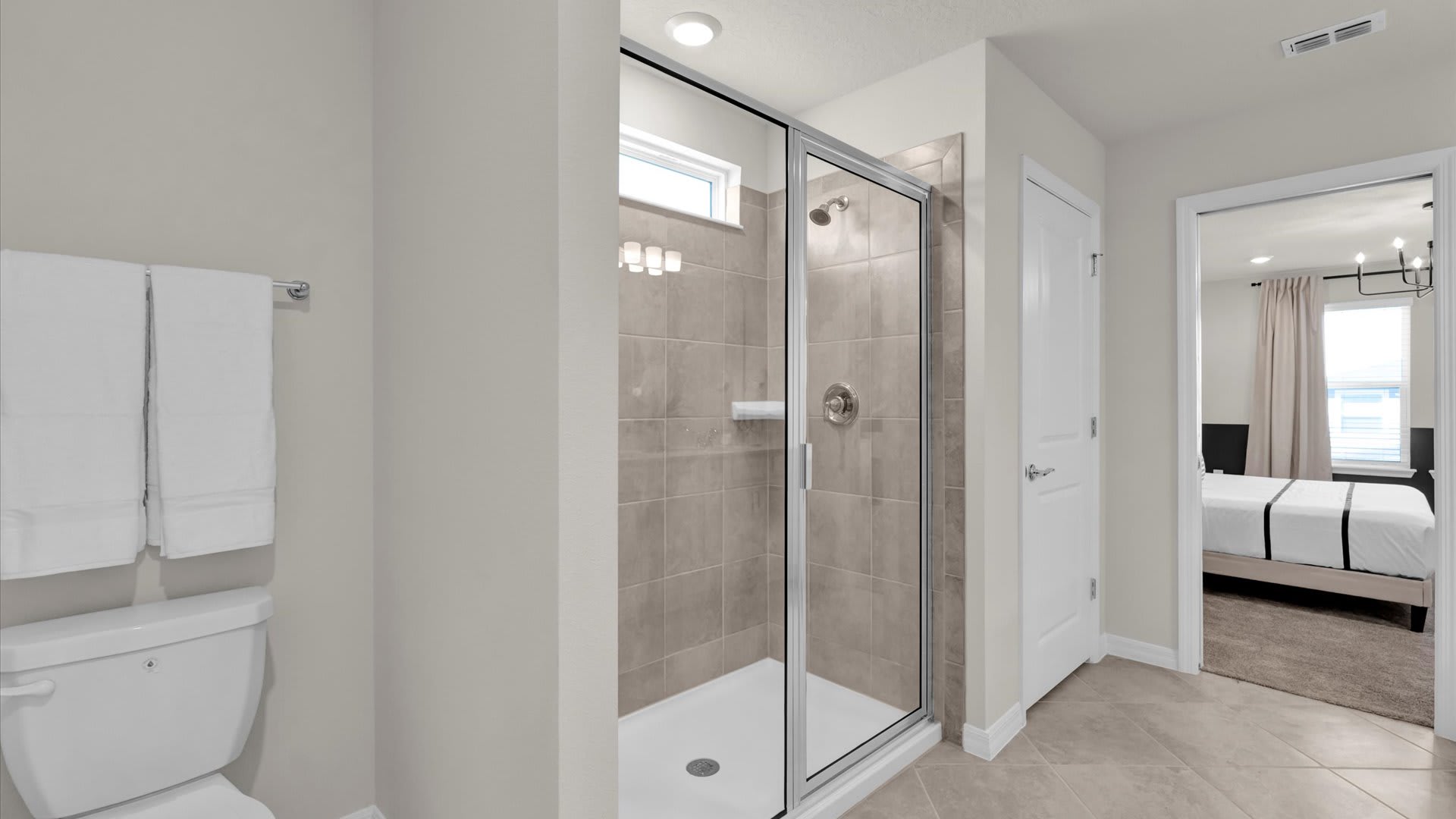 View of the bathroom's walk-in shower, enclosed by glass and featuring light brown subway tiles. A toilet is visible to the left, and a white door leads to the adjacent bedroom.